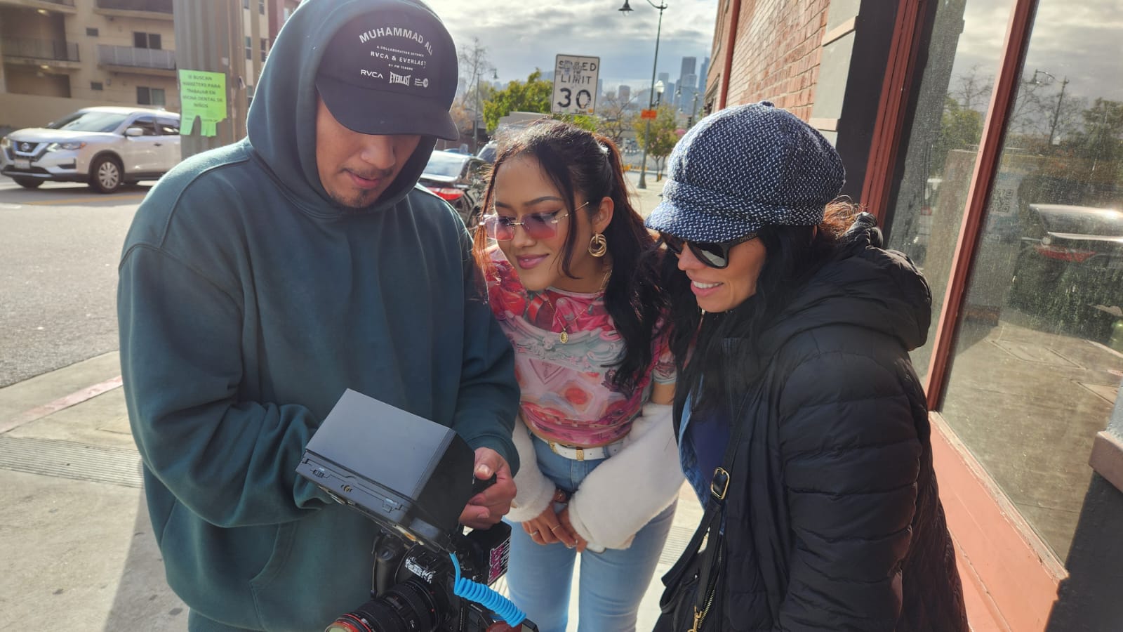 Three people looking at a camera outside a building, with cars and a street in the background.