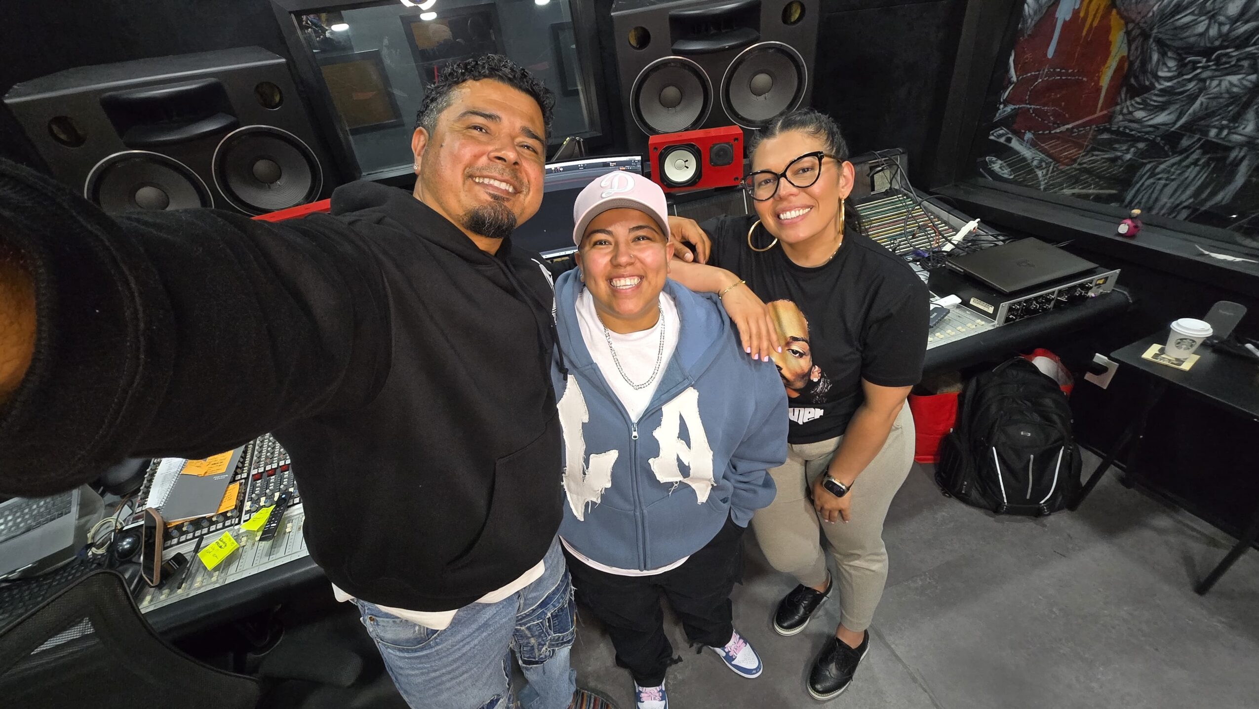 Three people smiling in a recording studio, taking a selfie together, with audio equipment and speakers in the background.