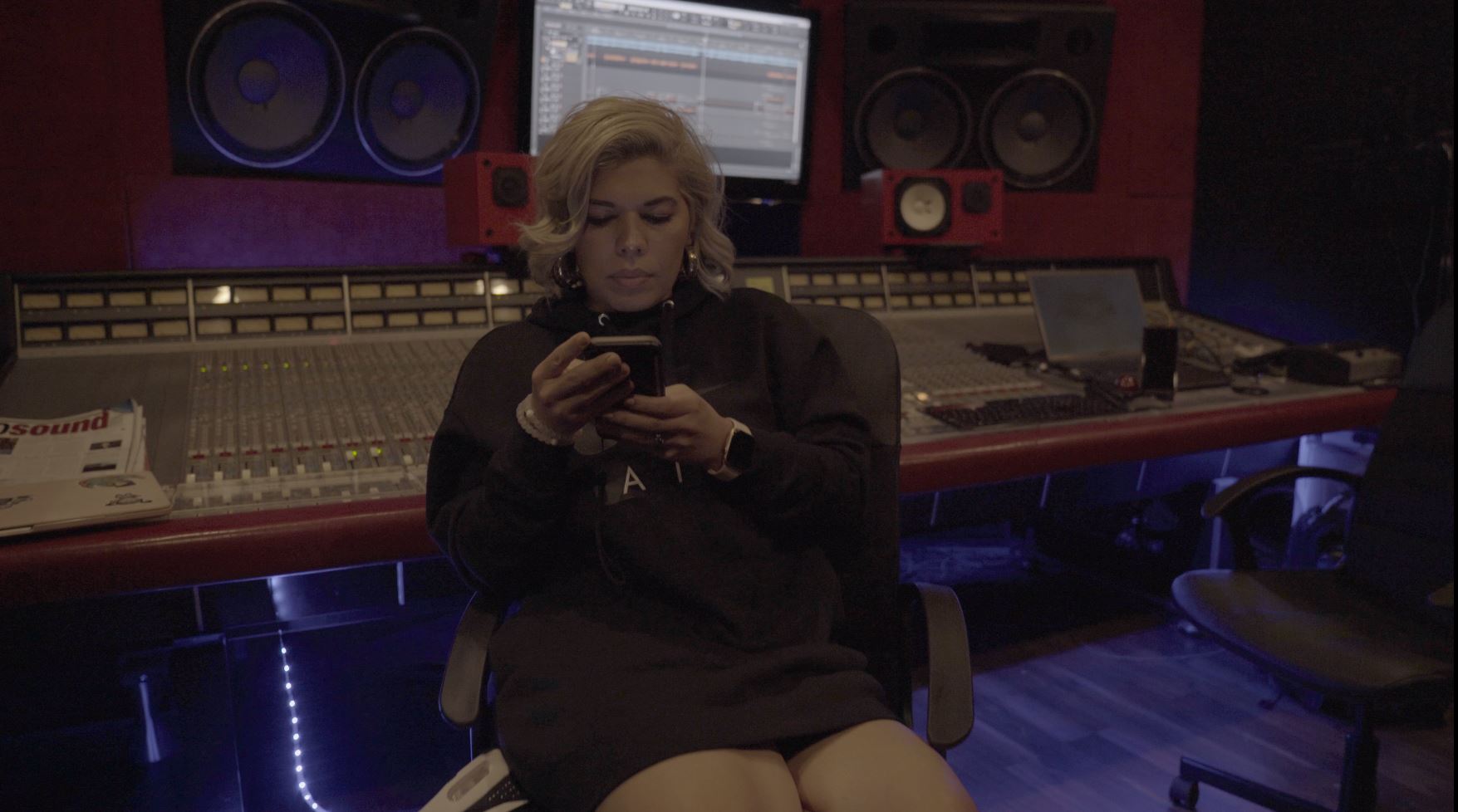 Person sitting in a recording studio, looking at a phone, with audio equipment and computer screens behind them.