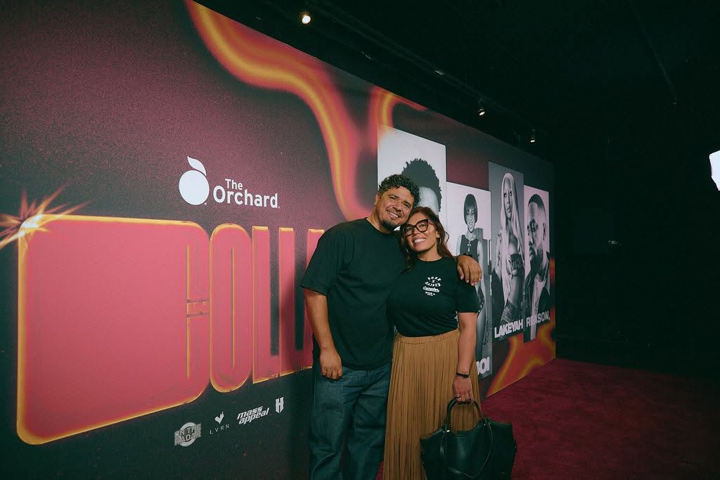 Two people standing together in front of a large event poster, smiling, with a dark background and red carpet.