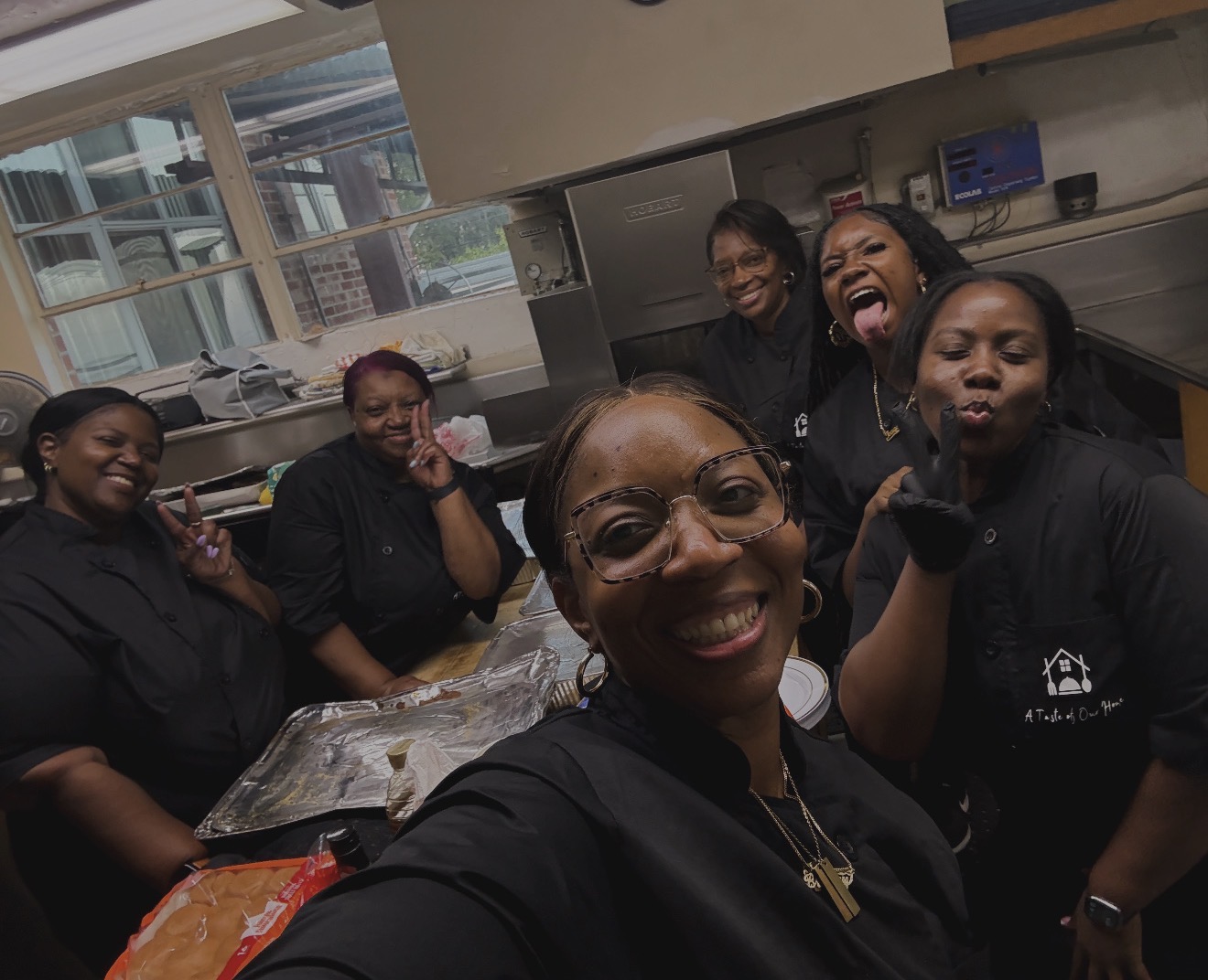 Group of six women smiling and posing in a kitchen, some making playful gestures, with a window and kitchen appliances in the background.