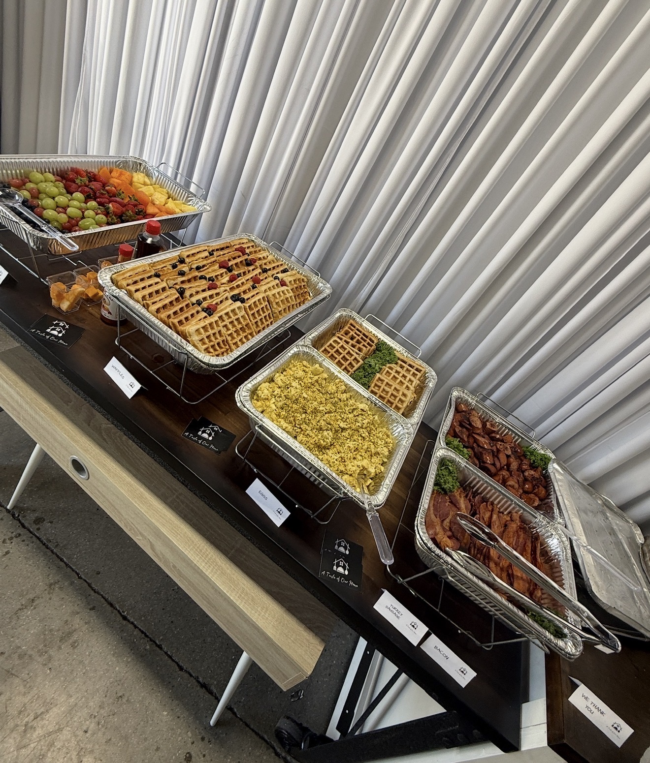 Assorted food items including fruit, waffles, and cooked dishes arranged on a black table with a silver curtain background.