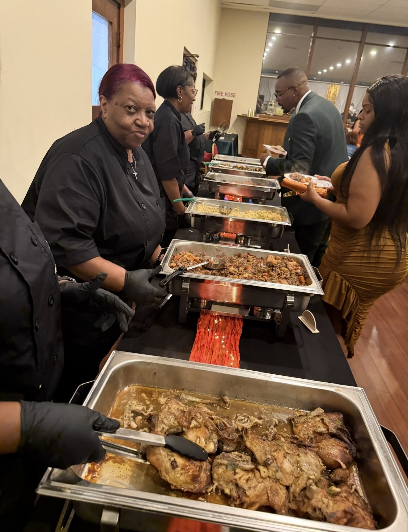 People serving food at a buffet table indoors, with trays of cooked meat and side dishes, and guests selecting food.
