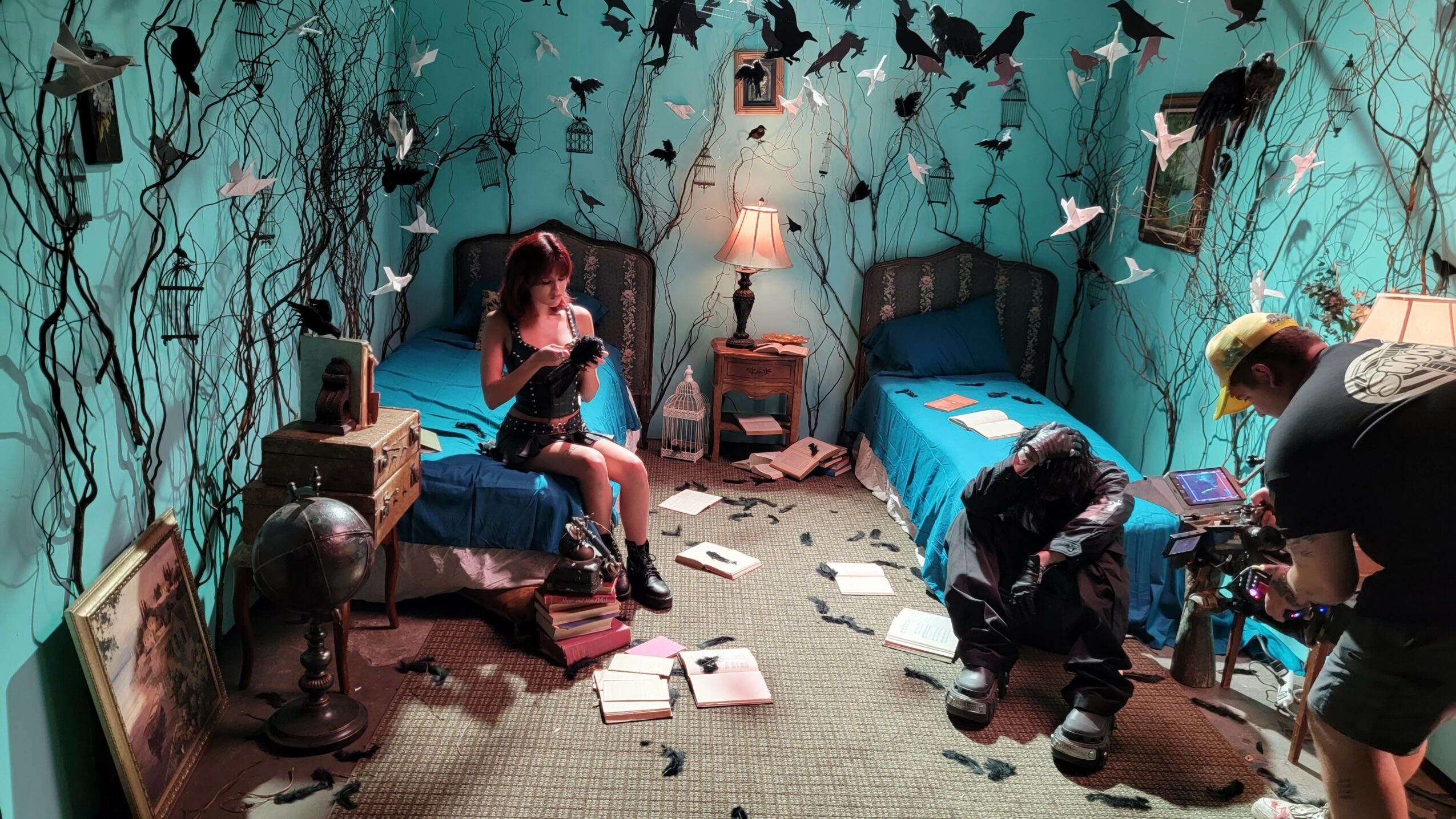 Room with two beds, blue walls, and paper birds hanging, cluttered with books, shoes, and a person reading.