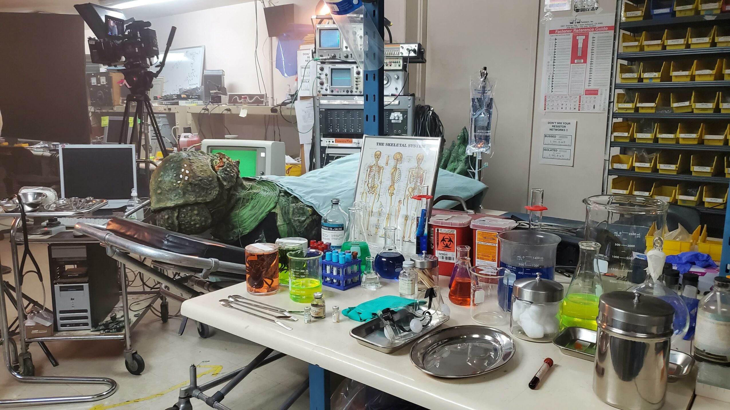 Cluttered workspace with equipment, supplies, and a camera on a tripod, in a room with shelves and electronic devices.