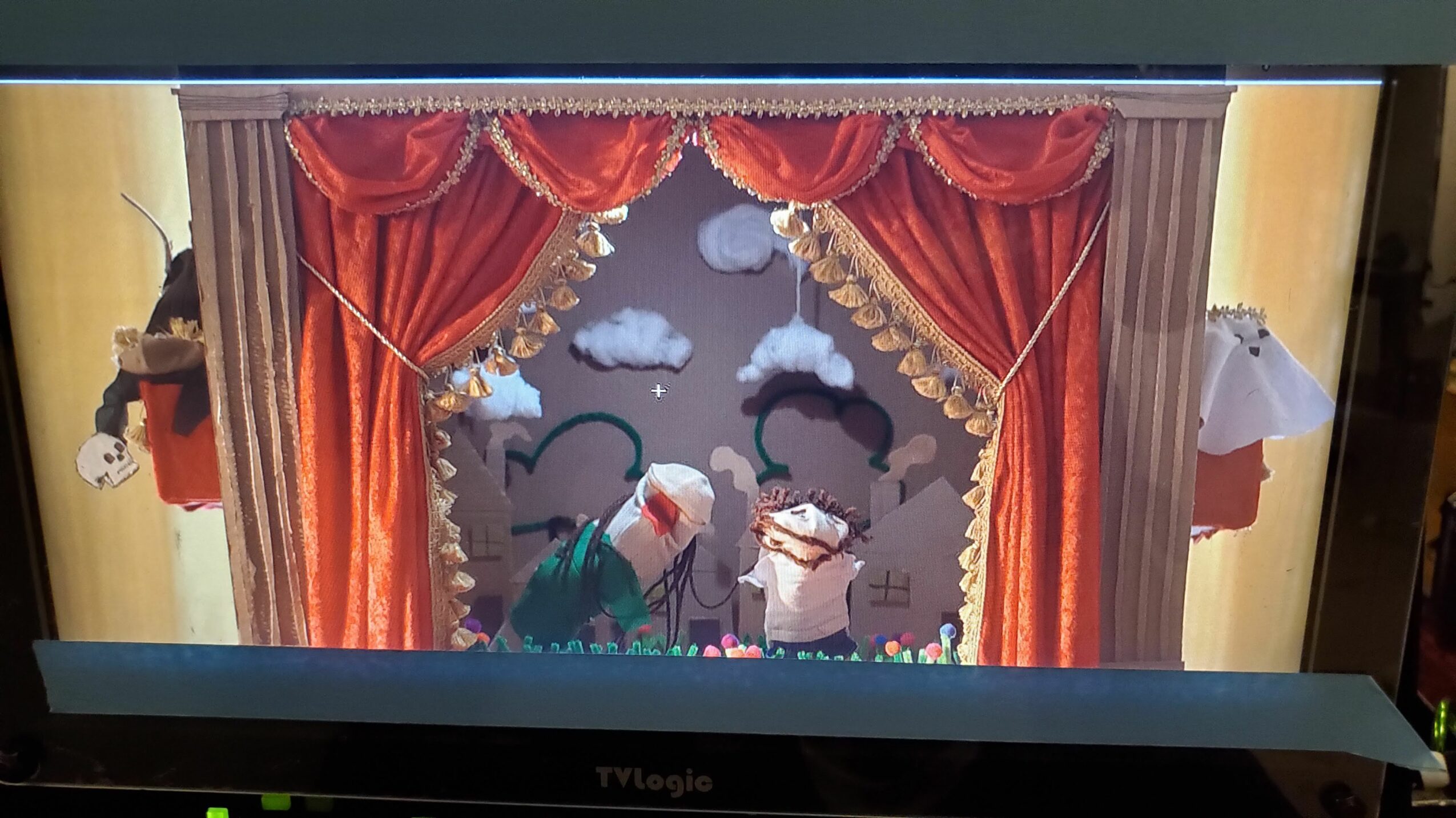Two puppets on a stage with red curtains and a cloudy sky backdrop, performing a scene.