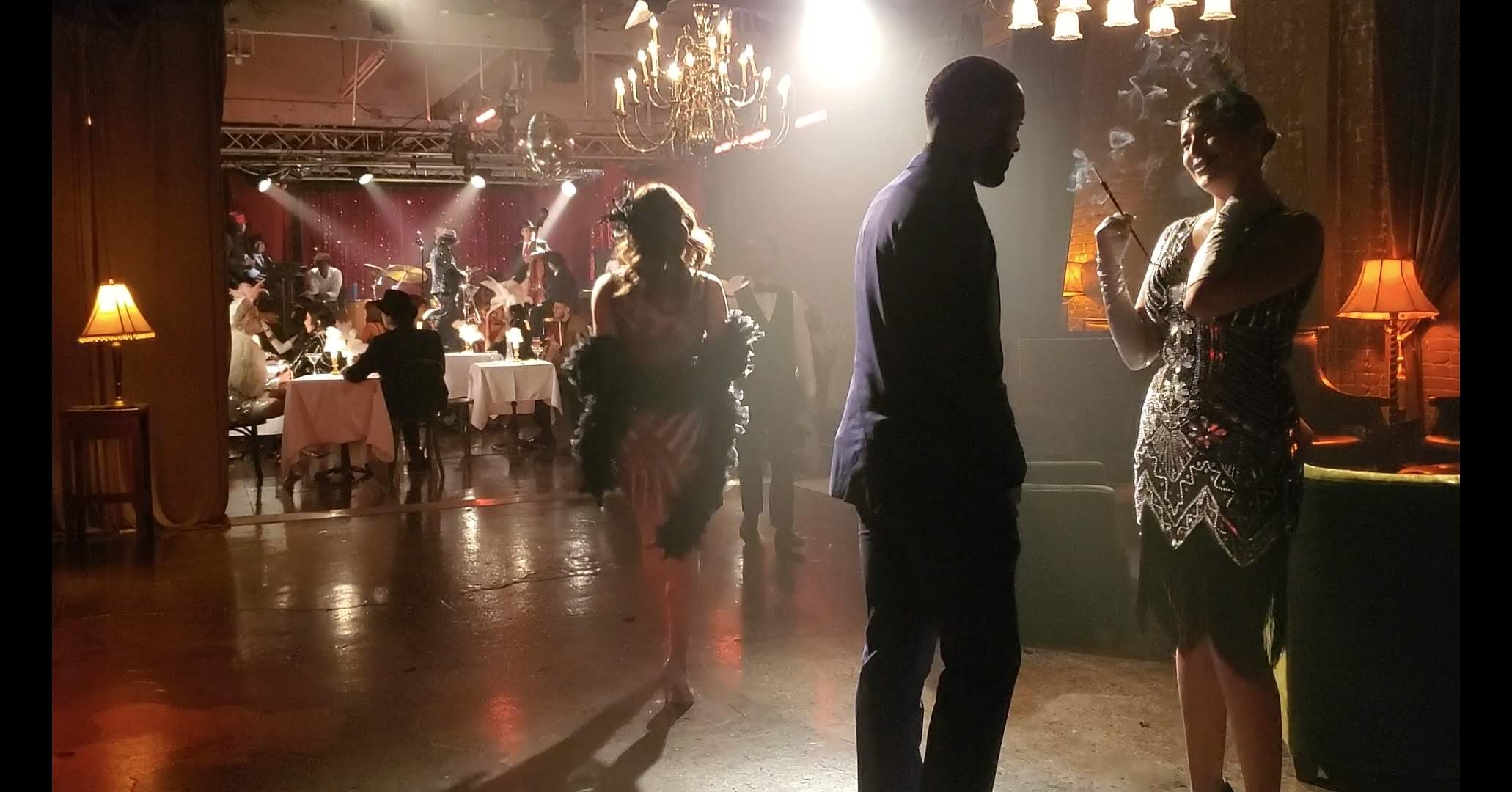 People dancing and socializing in a dimly lit, elegant event space with chandeliers and warm lighting.