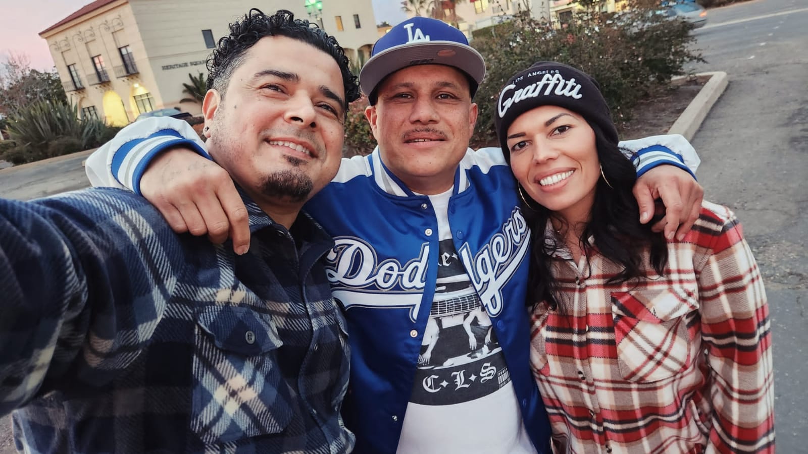 Three people smiling outdoors, arms around each other, wearing casual clothing and hats, with buildings and trees in the background.