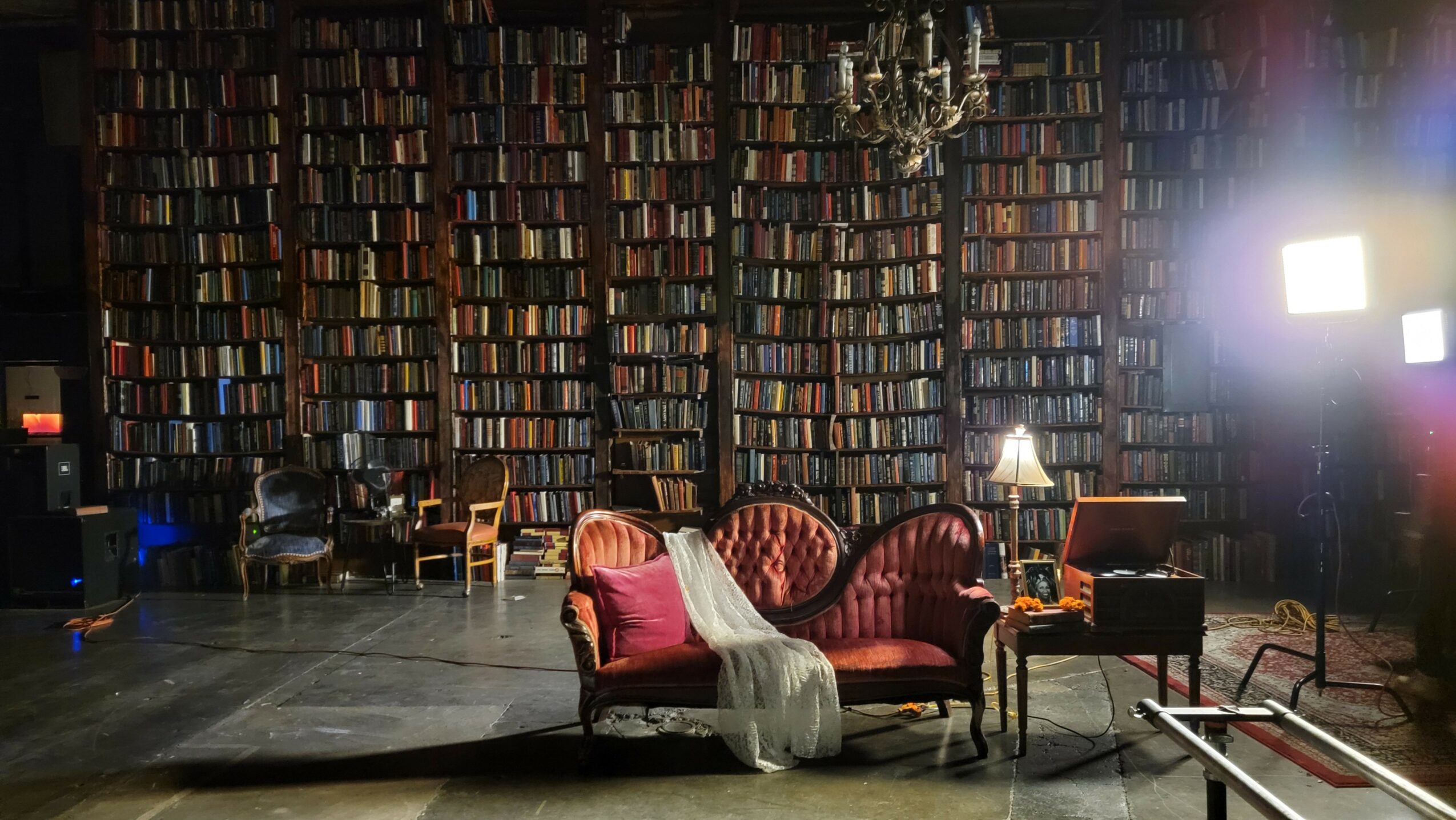 Stage with a pink sofa, a person in a white dress, and a library backdrop of bookshelves, with stage lighting.