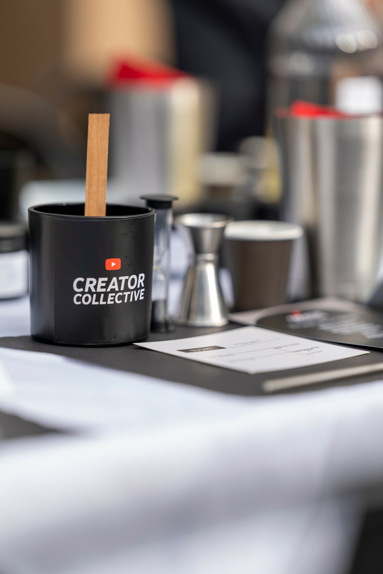 Black cup with wooden stir stick and logo, surrounded by metal containers and a white tablecloth.