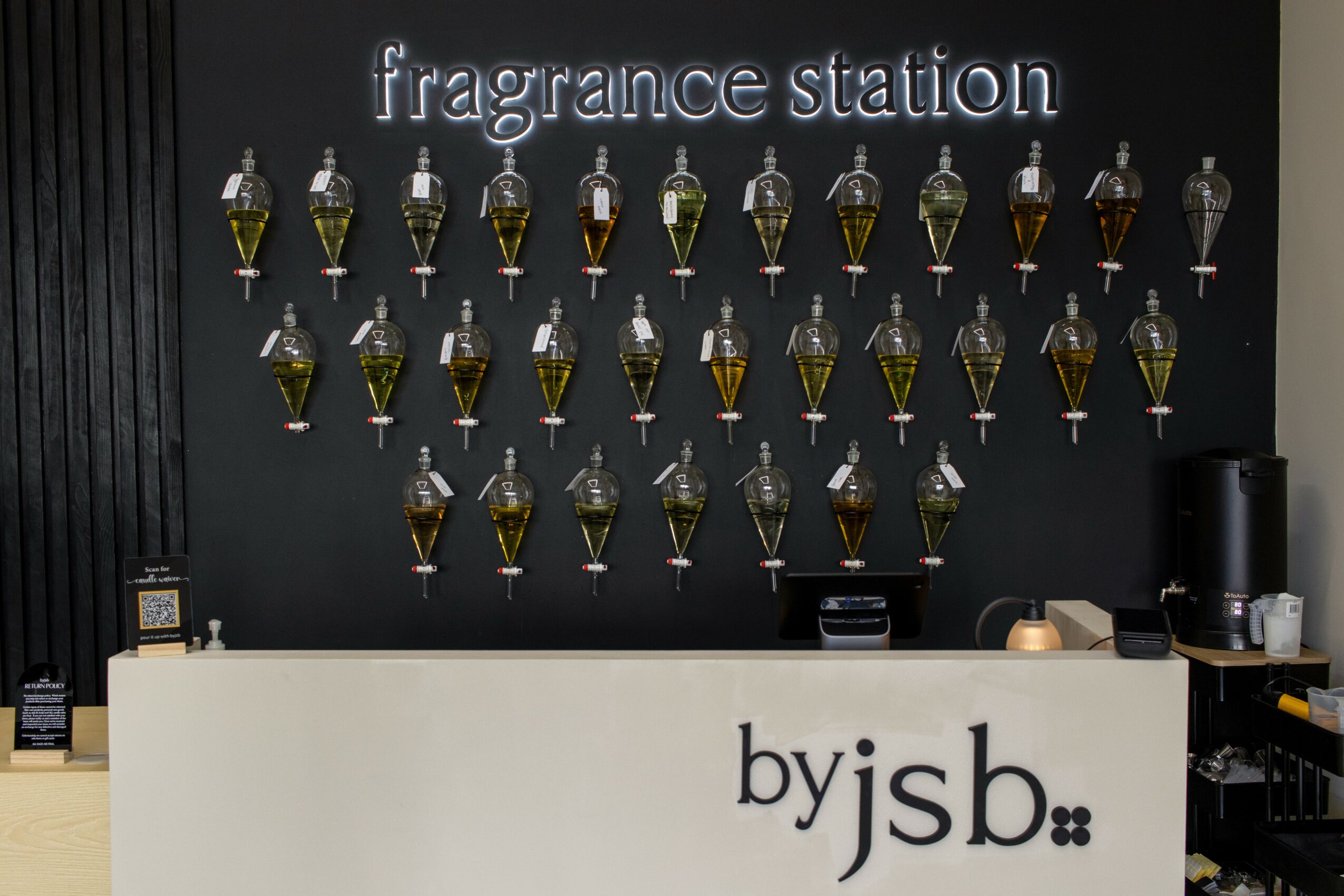 Display of fragrance bottles on a black wall with a white counter in front, labeled 'by jsb'.