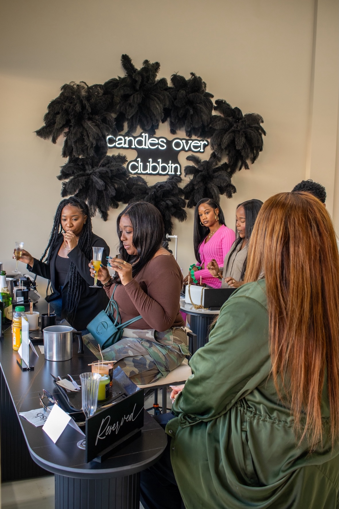 Group of women at a candle-making event, some holding drinks, with a decorative black foliage backdrop and a sign reading 'candles over clubbin'.