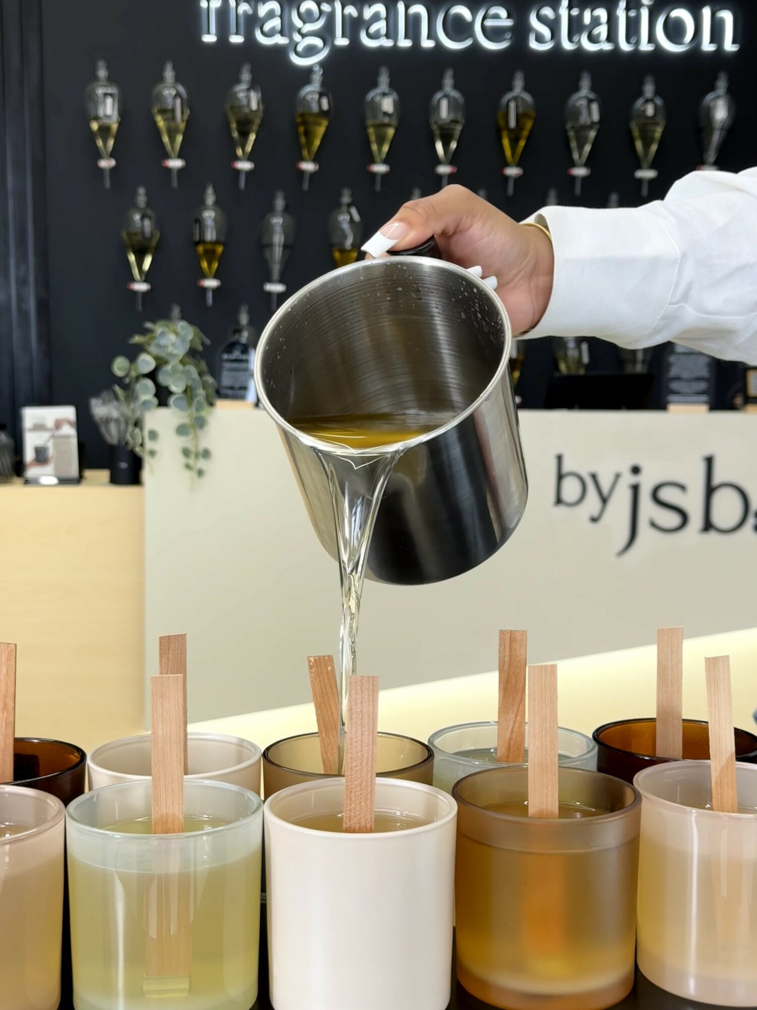 Person pouring liquid into a white cup at a fragrance station, with multiple cups and fragrance bottles in background.