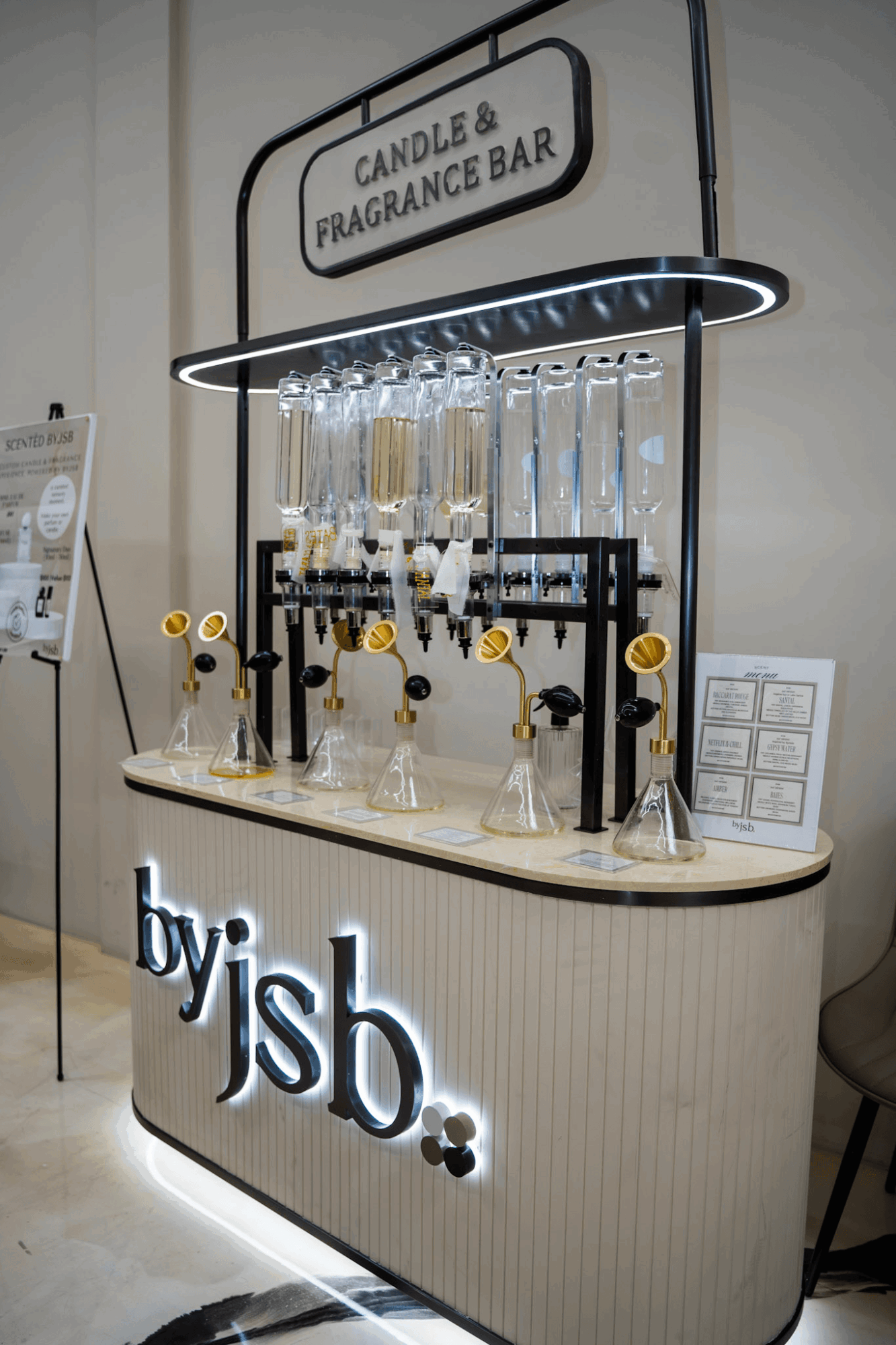 Display with glass bottles, a sign reading 'CANDLE & FRAGRANCE BAR,' and a lit sign 'by jsb.'