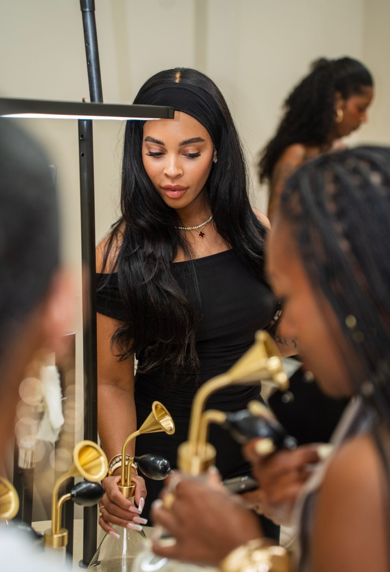 Young woman with long dark hair in a black top, looking down, surrounded by people, in a room with gold fixtures.