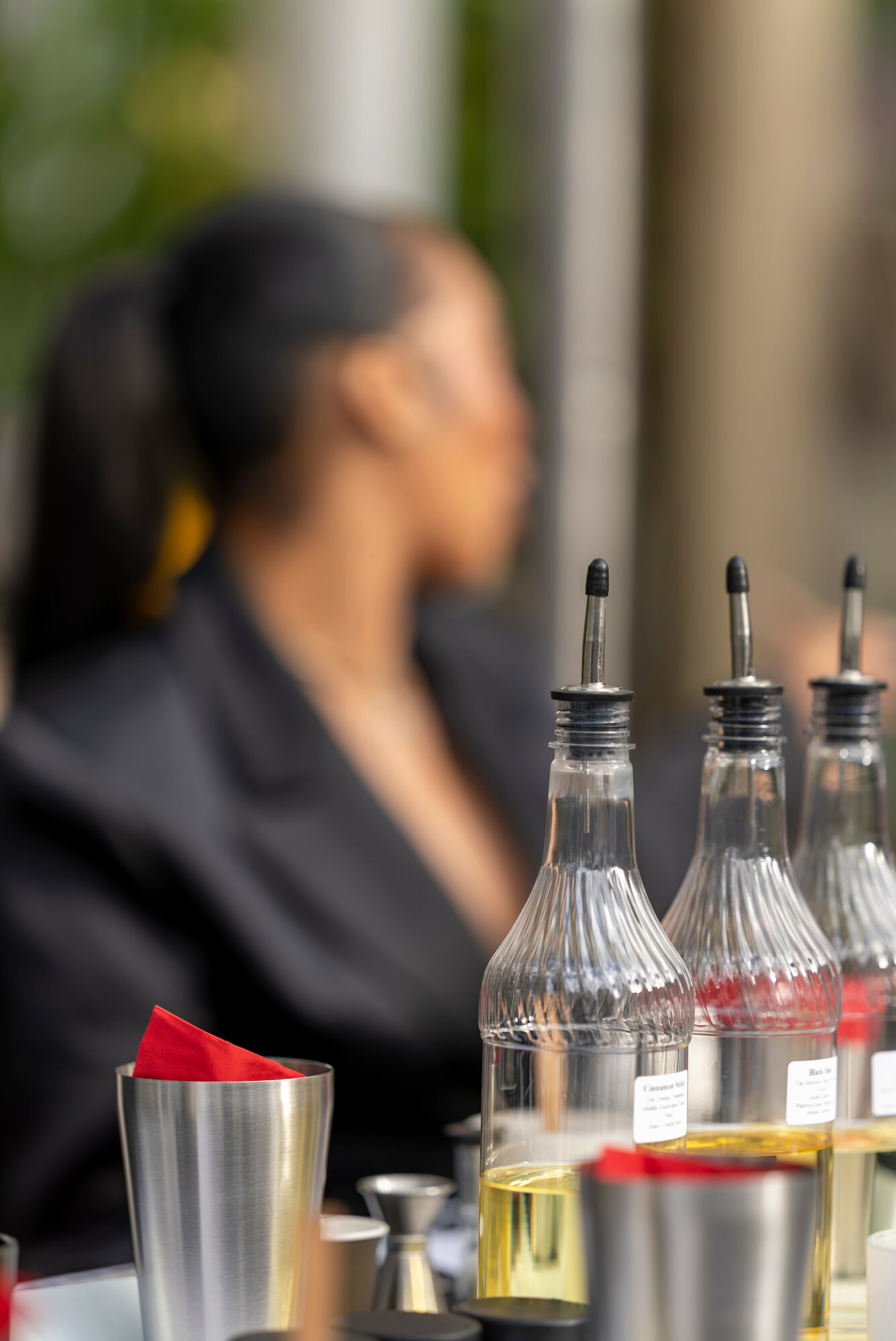 Blurred woman with dark hair tied back, sitting at a table with bottles and glasses, outdoors setting.
