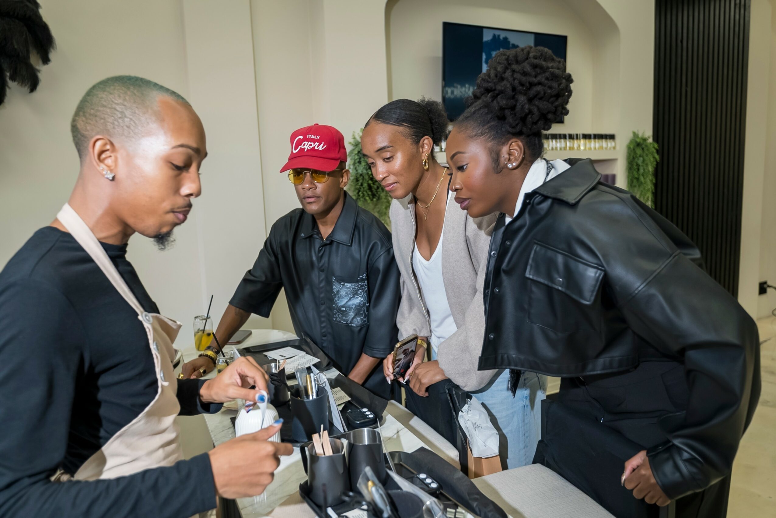 Four women gather around a table with makeup products, engaging in a discussion or demonstration in a room.