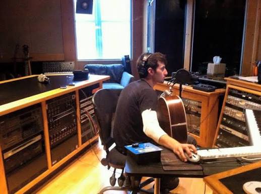Person sitting in a recording studio with a guitar, using a computer and audio equipment, window in background.