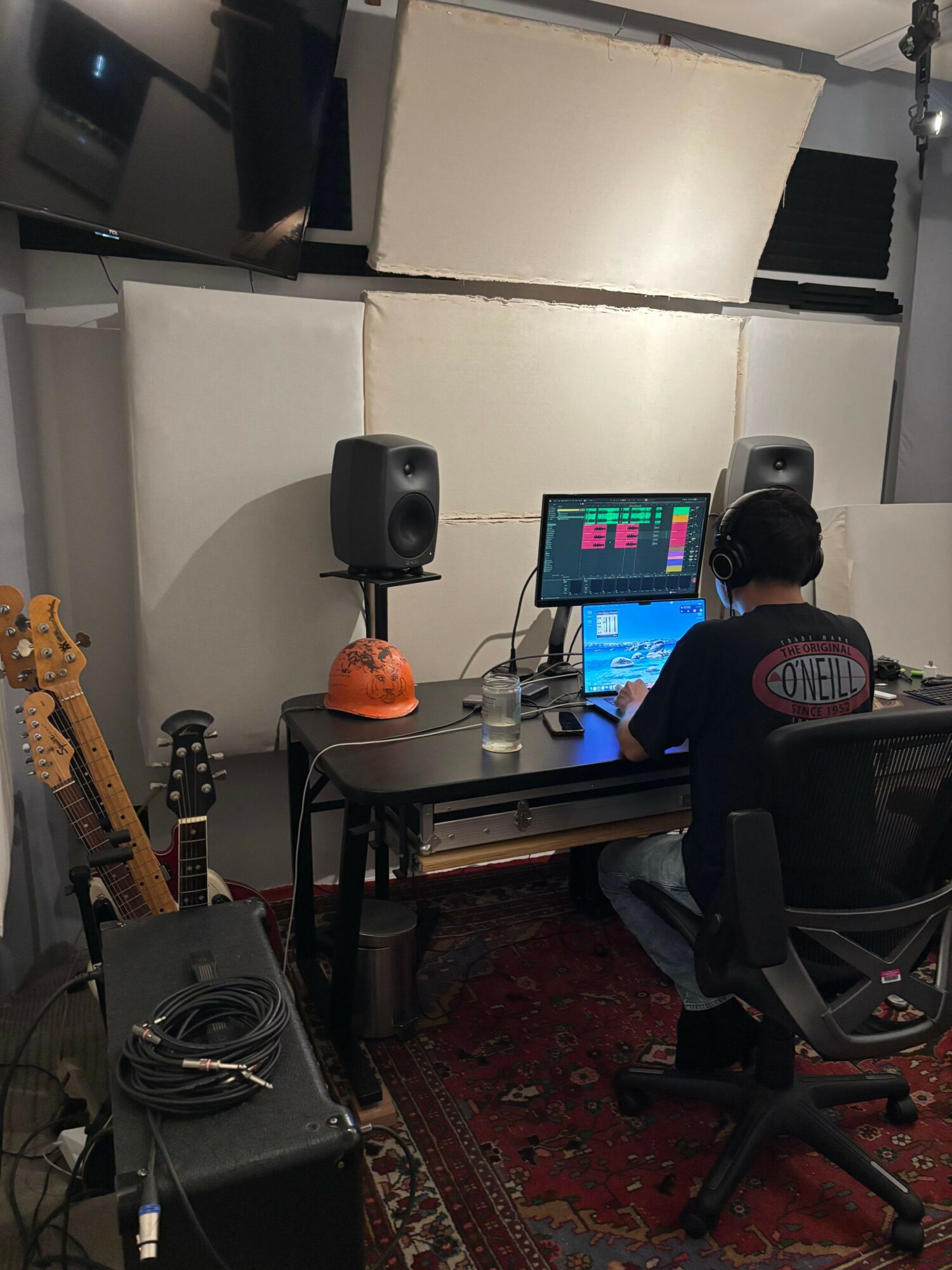 Person working at a computer desk with speakers, a guitar, and a monitor in a room with white walls and a red carpet.