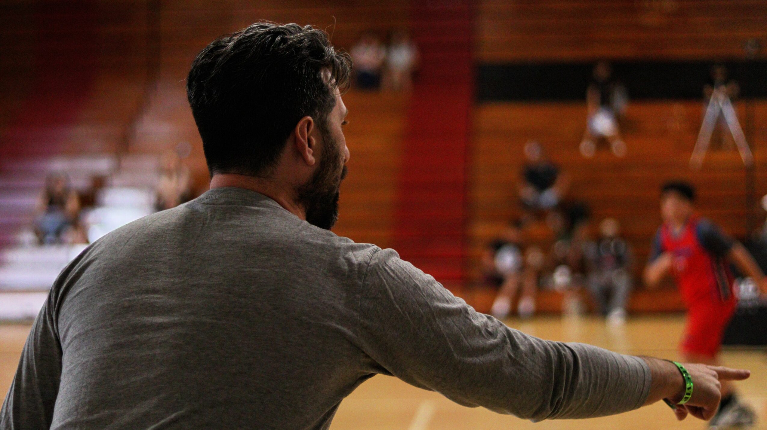 Man with dark hair and beard, wearing a gray shirt, extending his arm in a gymnasium with people in the background.