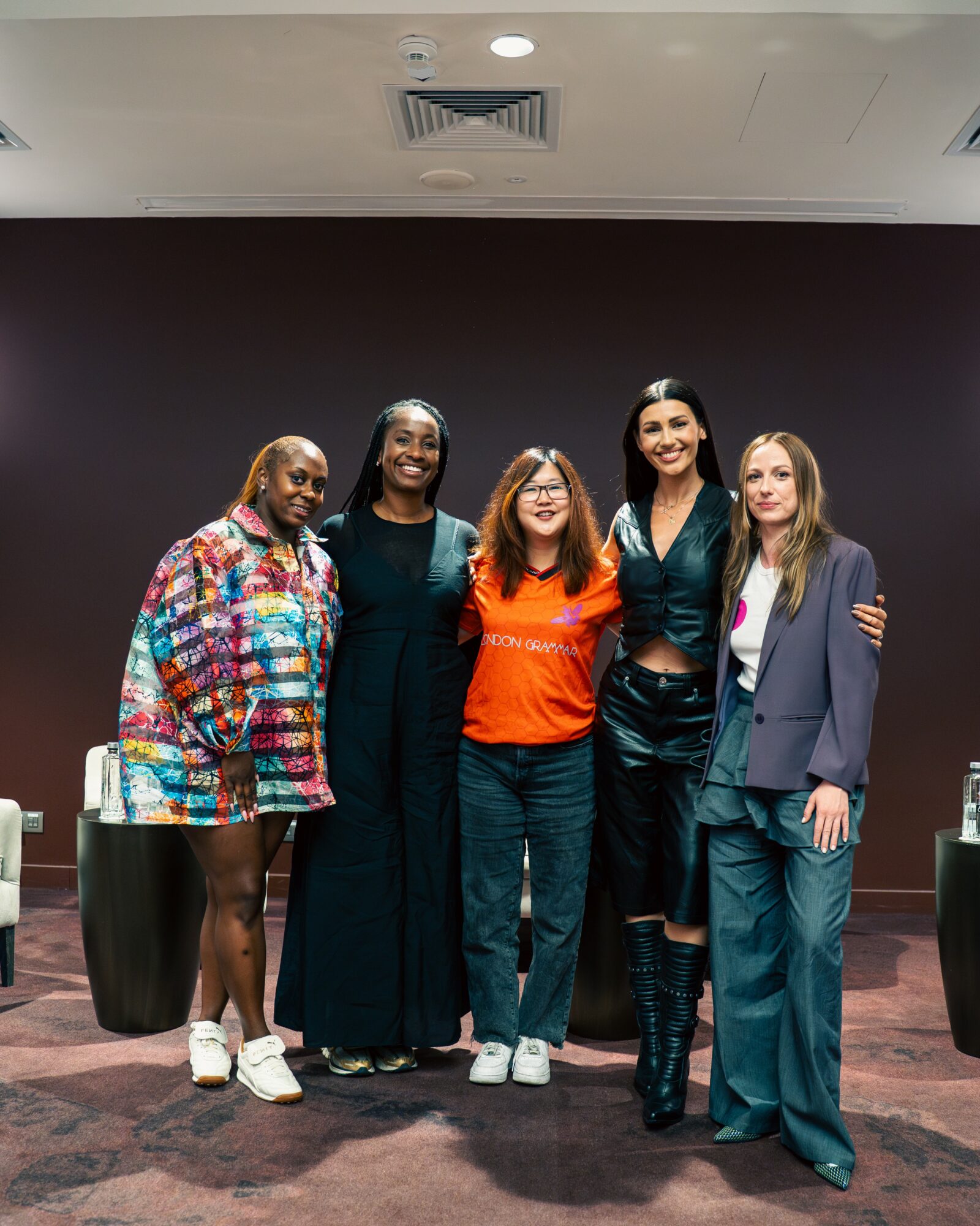 Five women standing together indoors, smiling, with a dark wall background and tables on each side.