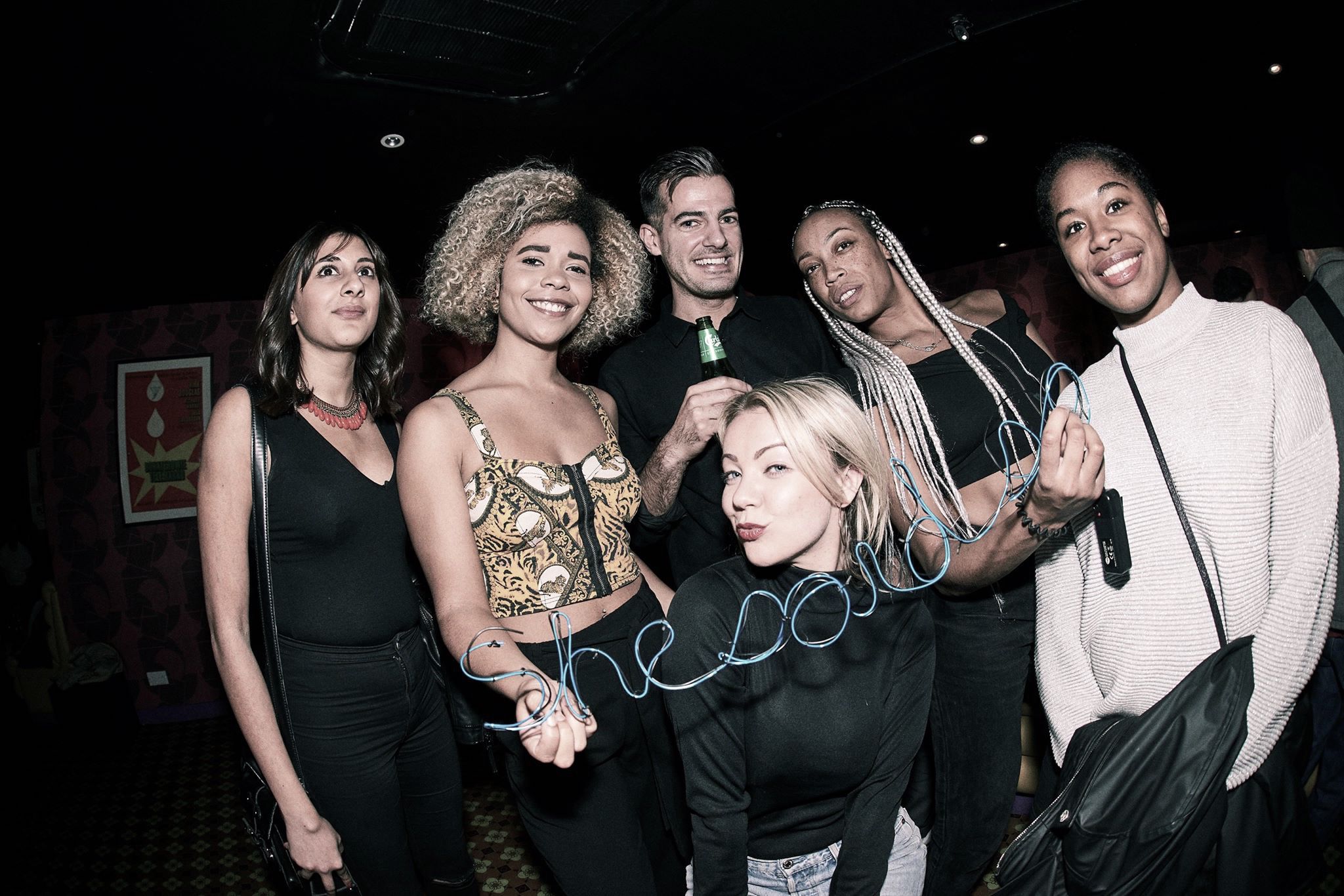 Group of six people smiling and posing together in a dark indoor setting.