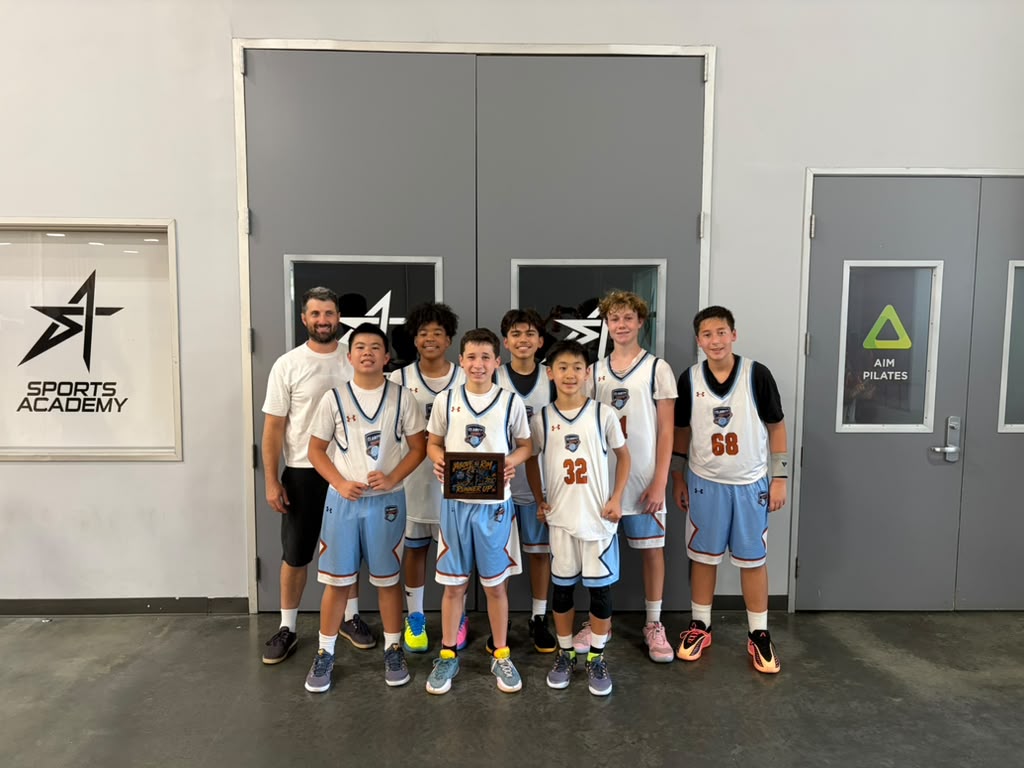 Group of young basketball players and coach standing in front of closed doors, holding a trophy, smiling.