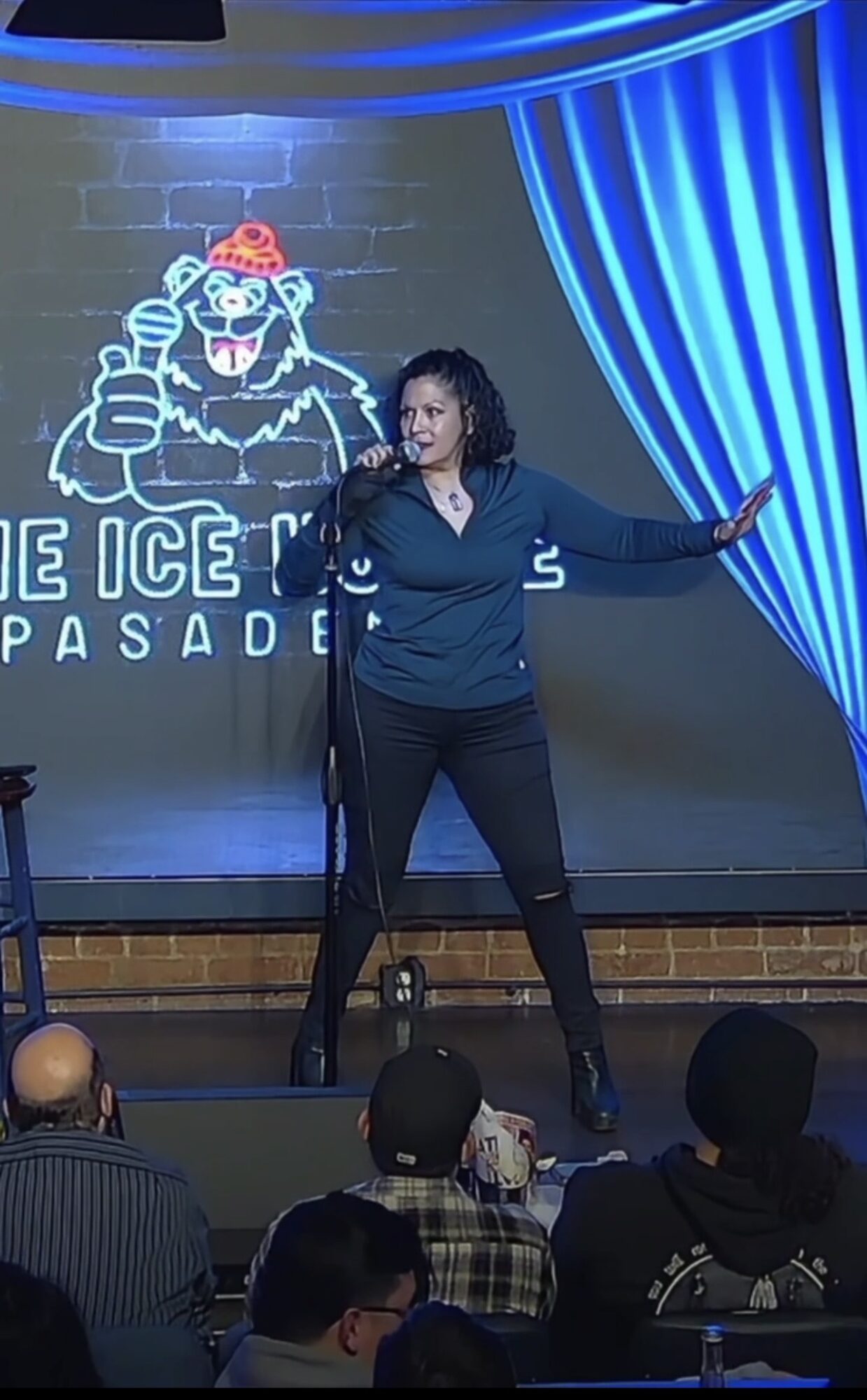 Woman standing on stage with a microphone, gesturing with her right hand, in front of a neon sign and blue curtains.