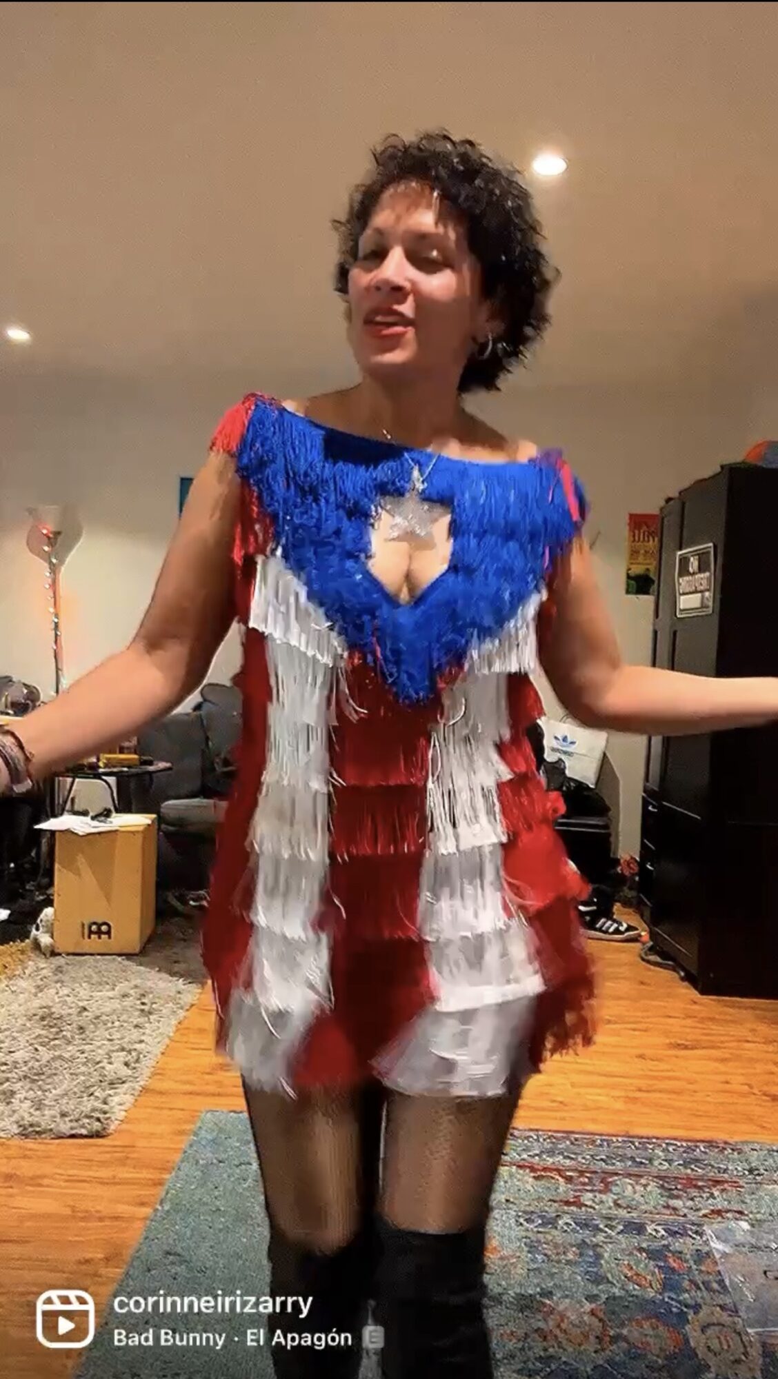 Person with curly hair wearing a colorful, fringed dress standing in a room with wooden floor and furniture.