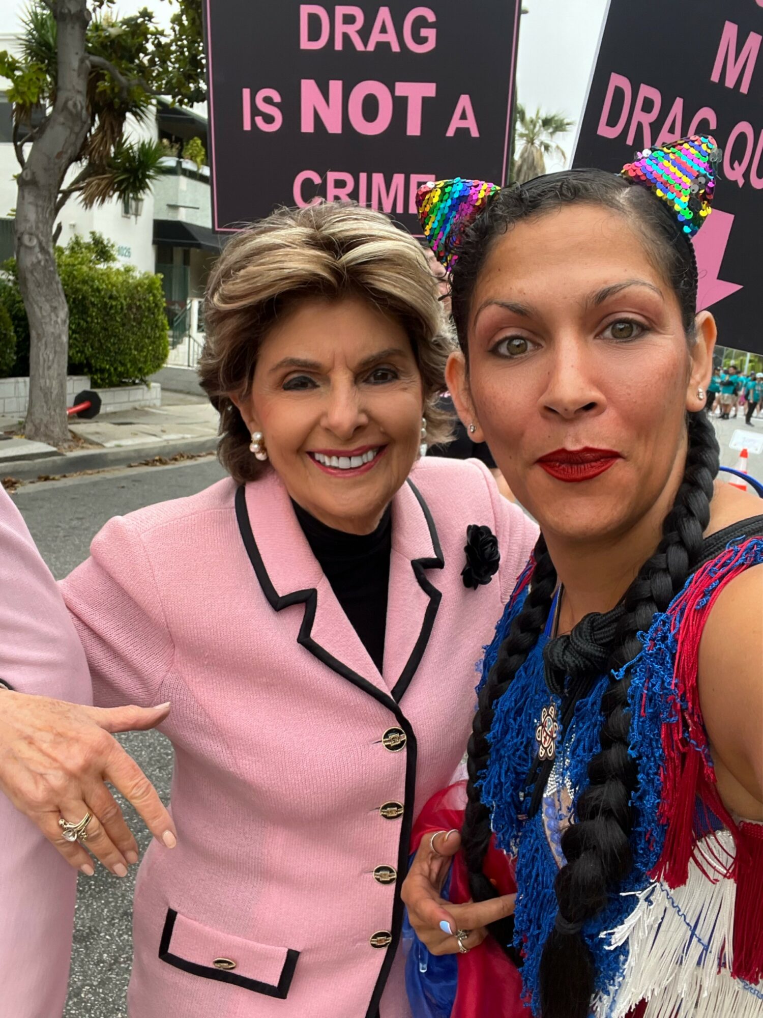 Two women smiling at a protest, holding signs that say 'Drag is NOT a crime,' outdoors with trees and buildings in background.