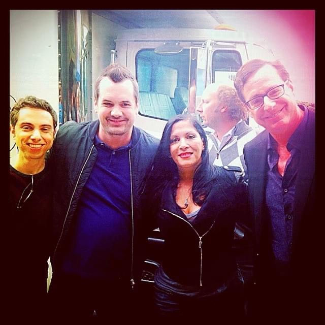 Four people standing together outdoors, smiling, with a vehicle and other people in the background.