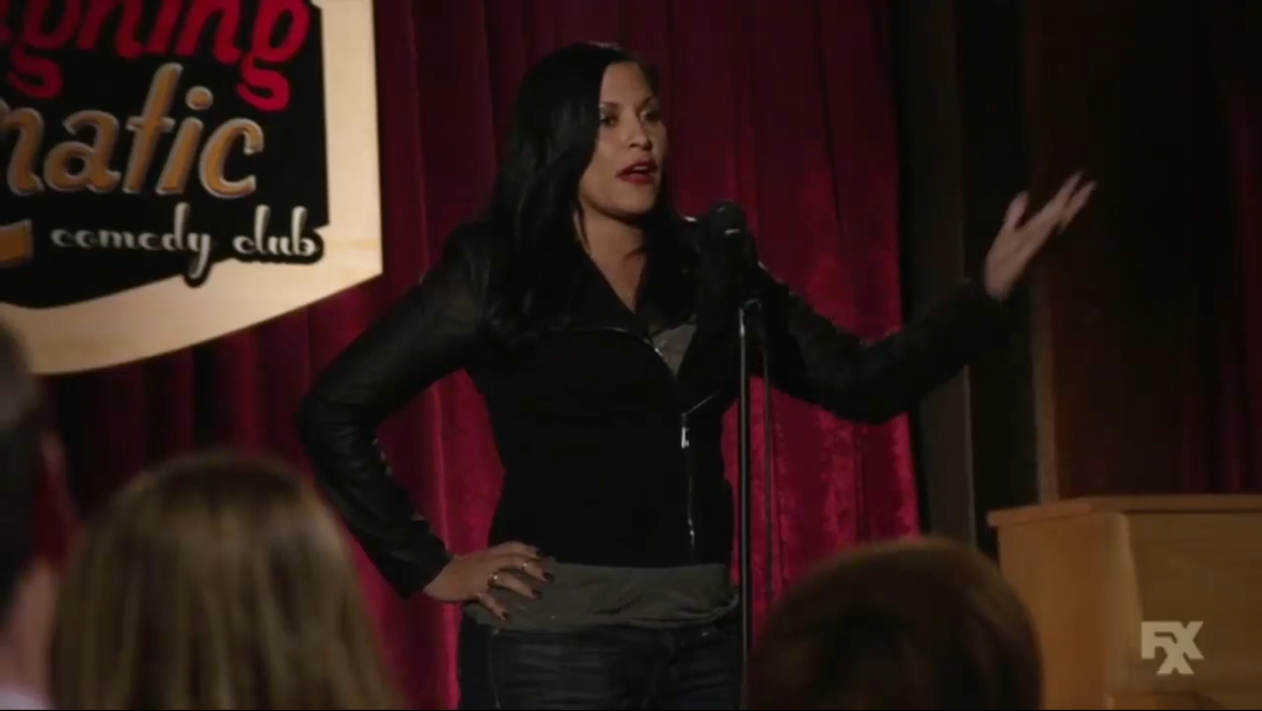 Woman with black hair and red lipstick speaking on stage with red curtains behind her.