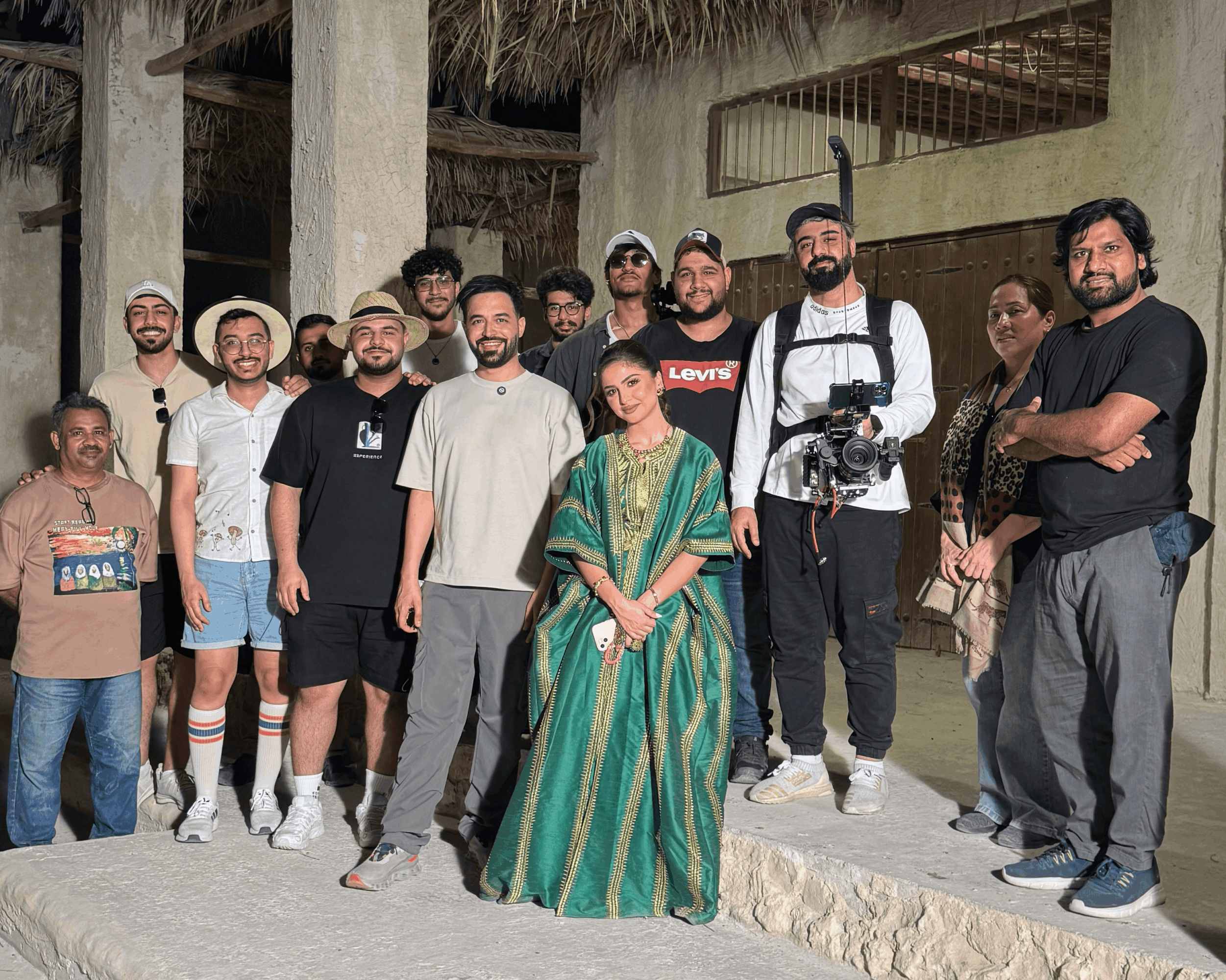 Group of people standing outdoors, some smiling, with a woman in a green sari in the center, sunlight, and rustic background.
