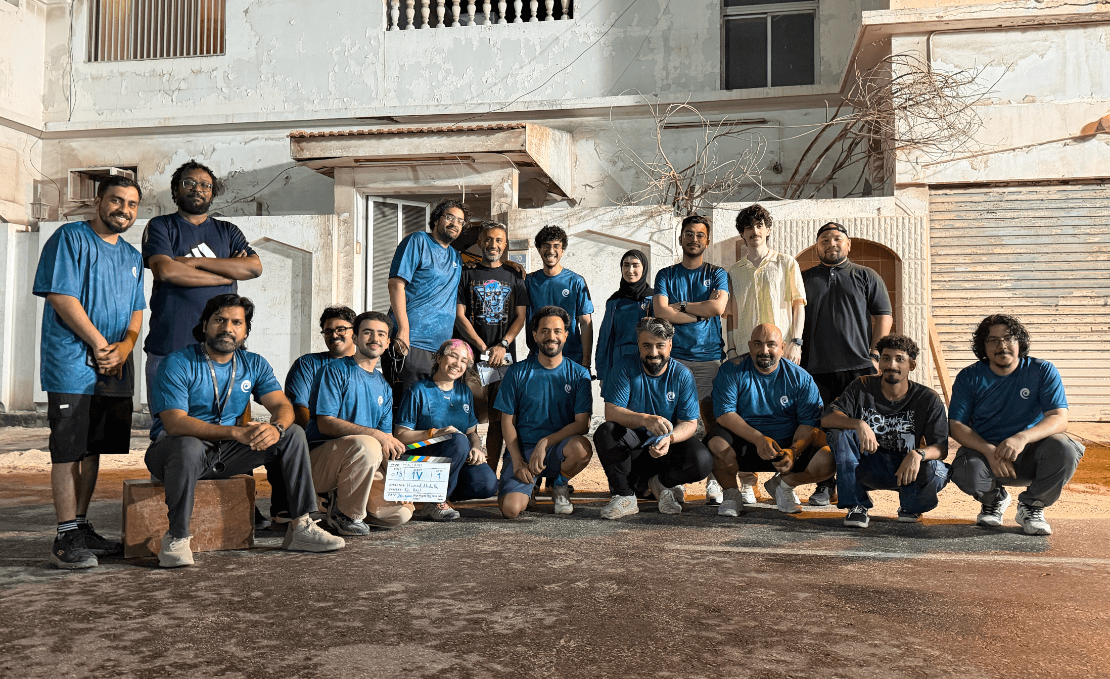 Group of people posing outdoors in front of a building, some standing and some kneeling, with a clapperboard, daytime.