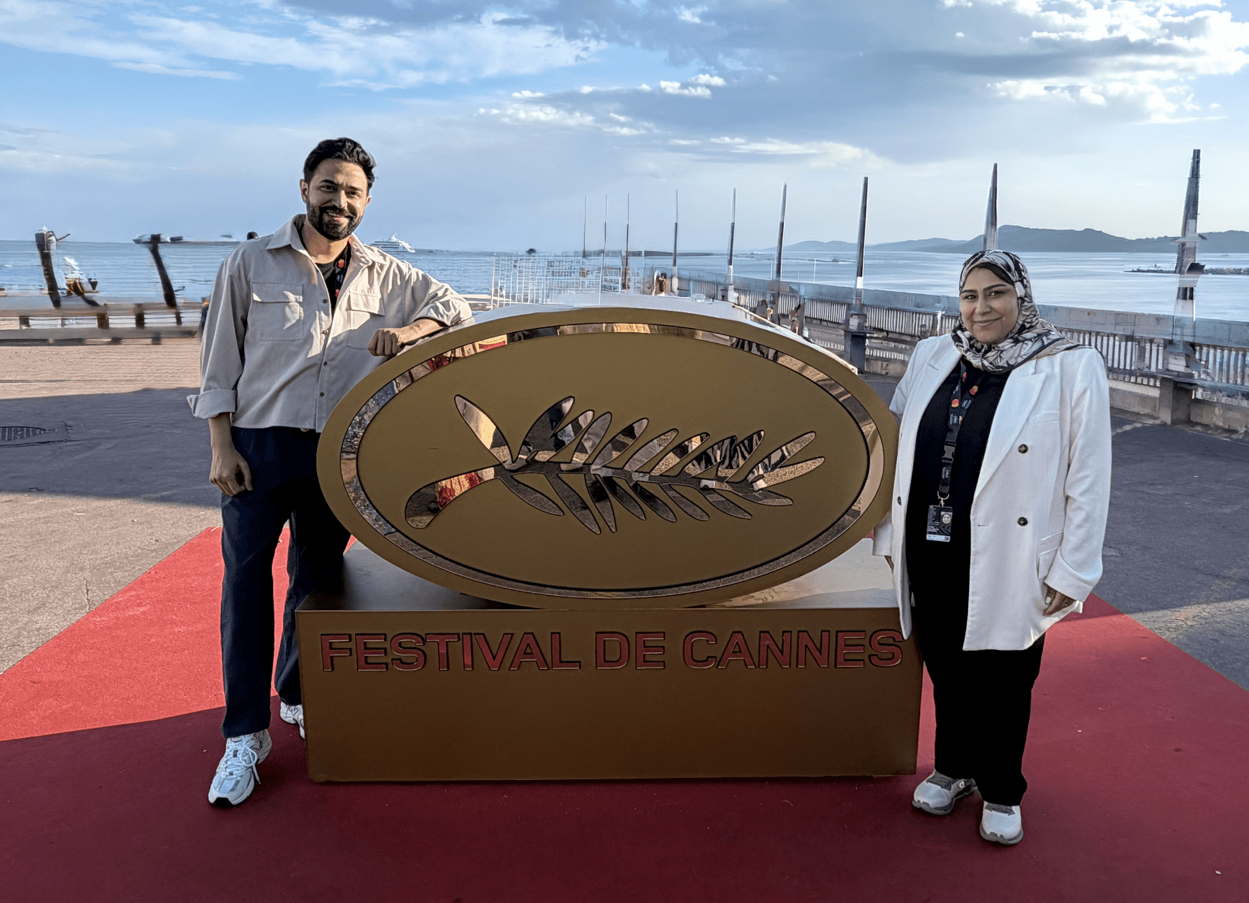 Two people stand outdoors near a large Cannes Film Festival sign, with a harbor and sailboats in the background.