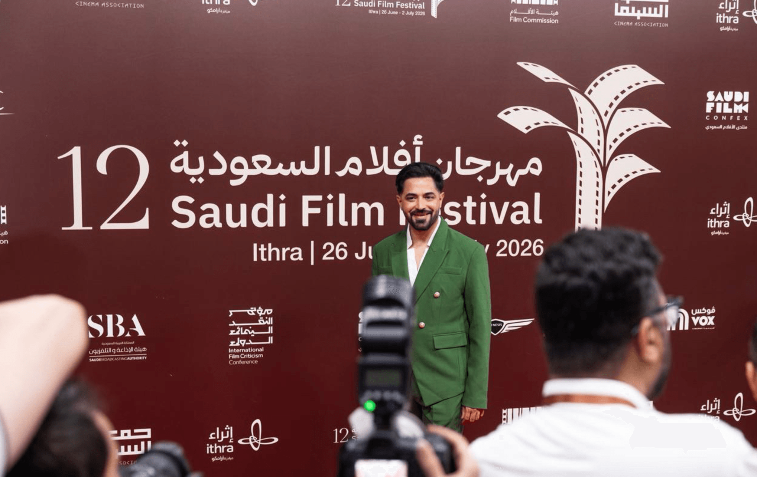 Man in green suit smiling at Saudi Film Festival event with a maroon backdrop and white text.