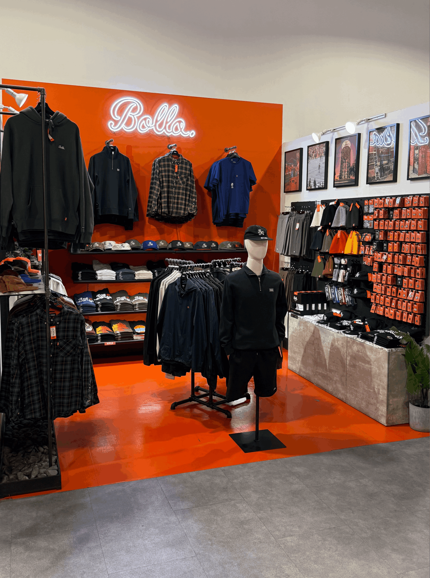Clothing store display with jackets, hats, and a mannequin, featuring a bright orange wall with a neon sign, and framed pictures on the side.