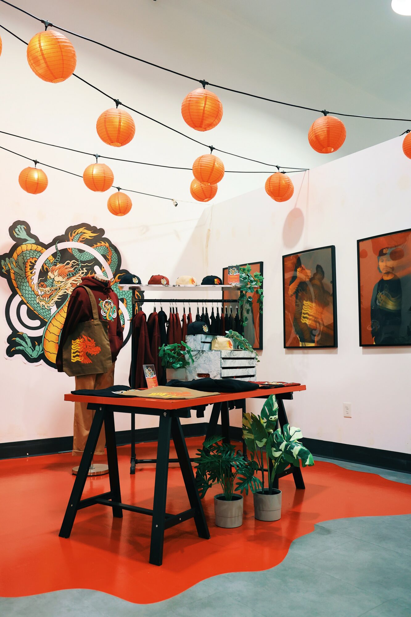 Art gallery with hanging orange lanterns, paintings on white walls, a table with potted plants, and colorful decor.