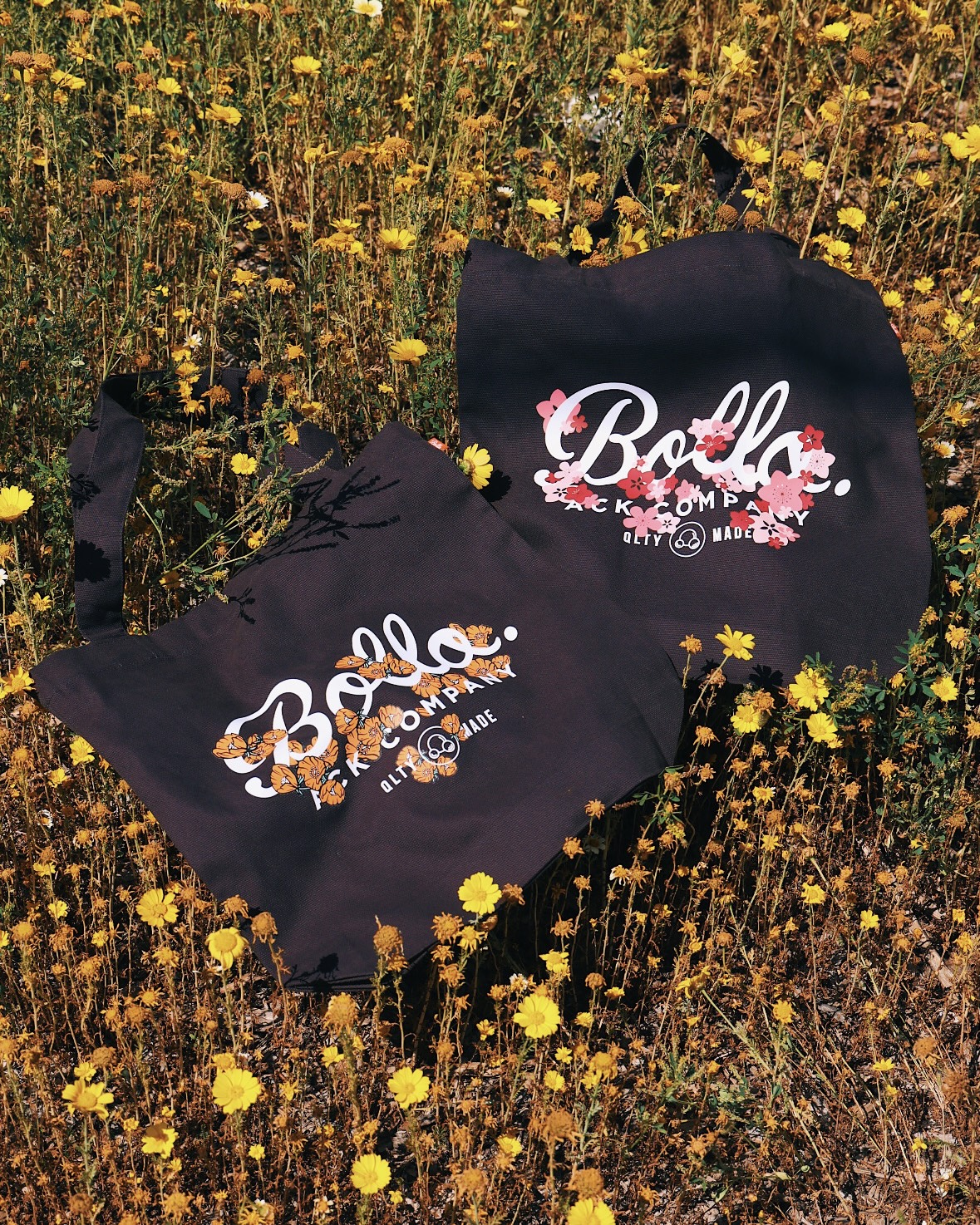 Two black bags with floral and text designs are placed among yellow wildflowers in a field.