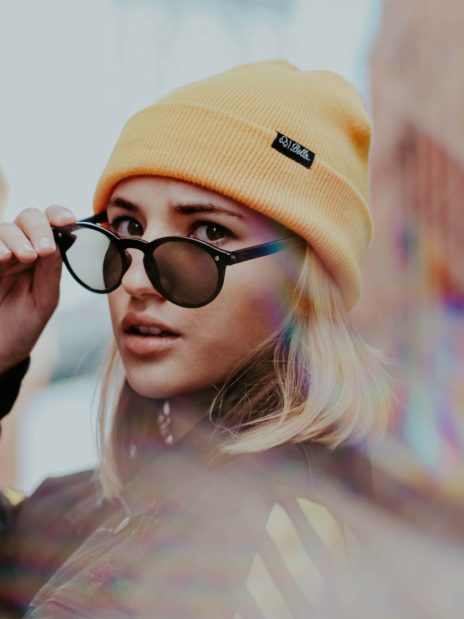 Young woman wearing a yellow beanie and sunglasses, looking over the glasses with a city background.