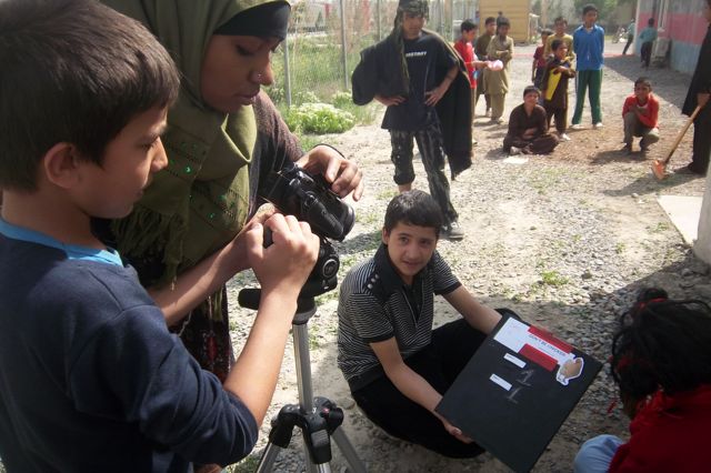Group of children and a woman outdoors, some sitting and some standing, with a camera and a display board. Twenty words.