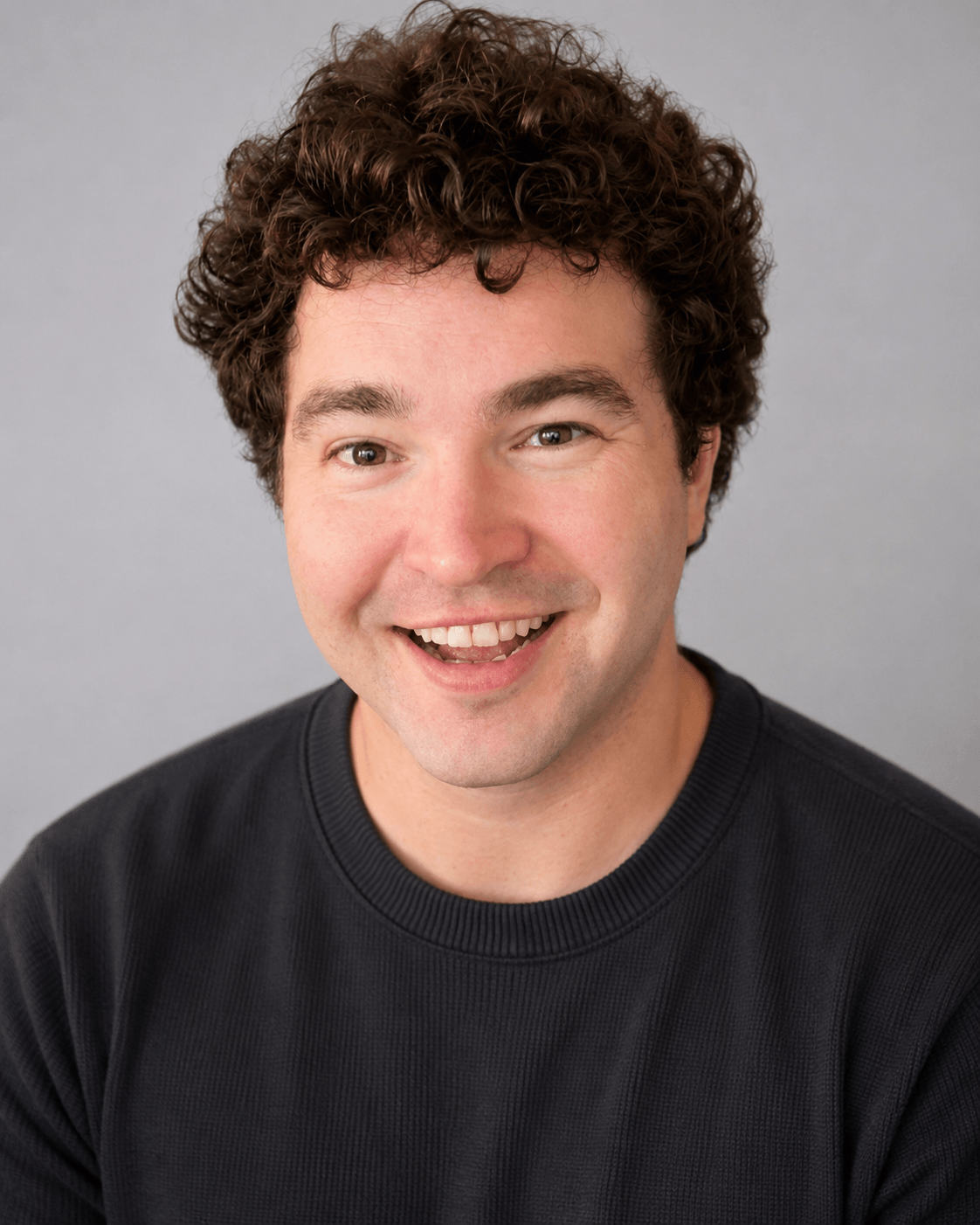 Smiling man with curly hair wearing a black shirt, facing forward, neutral background.