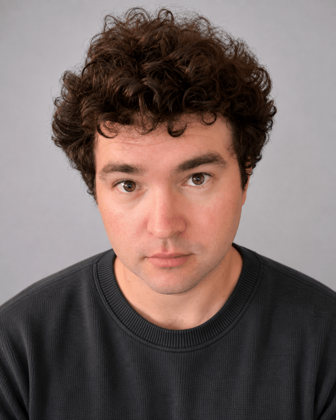 Young man with curly brown hair and light skin, wearing a black shirt, looking directly at the camera, neutral expression.