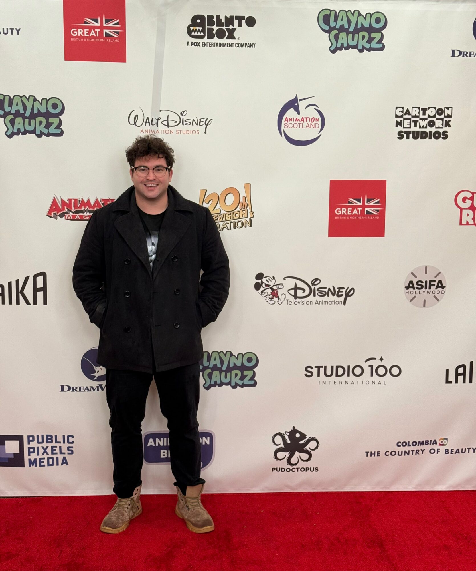 Young man with glasses and curly hair, wearing a black coat, standing on a red carpet in front of a white backdrop with logos.