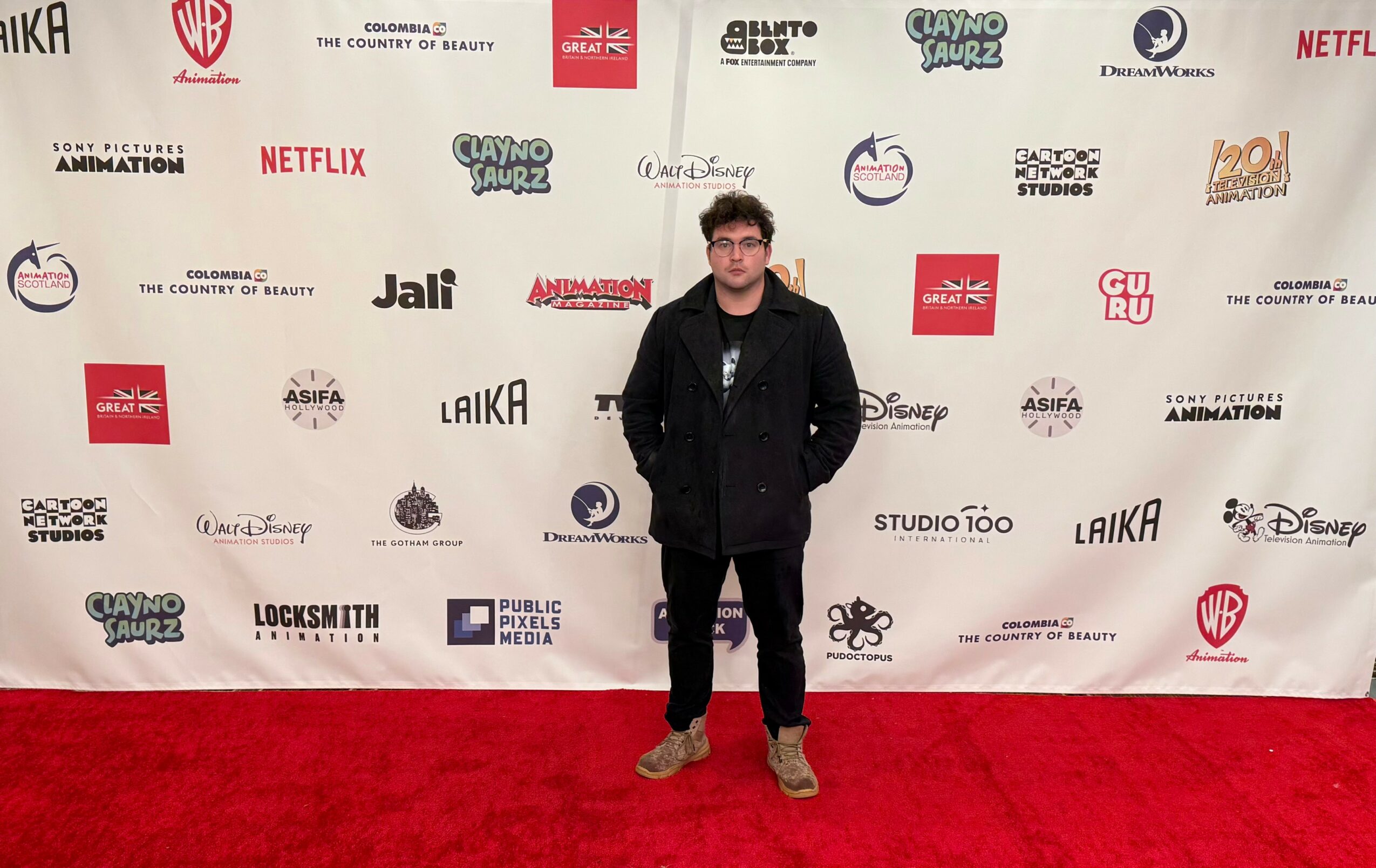Man standing on red carpet in front of a white backdrop with various logos, wearing glasses, black jacket, and brown shoes.