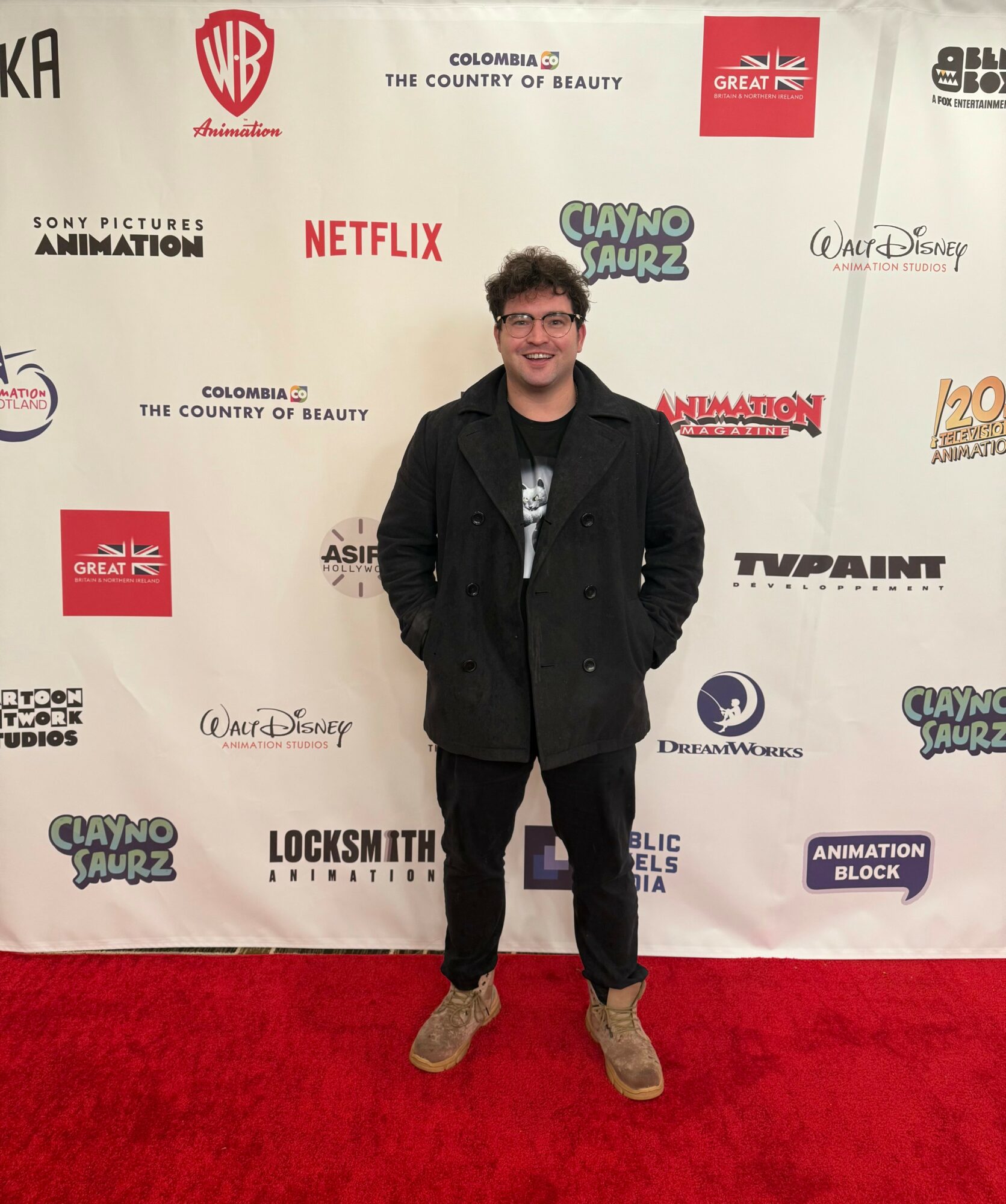 Young man with glasses and curly hair standing on red carpet in front of a white backdrop with logos, smiling, wearing a black coat and tan shoes.