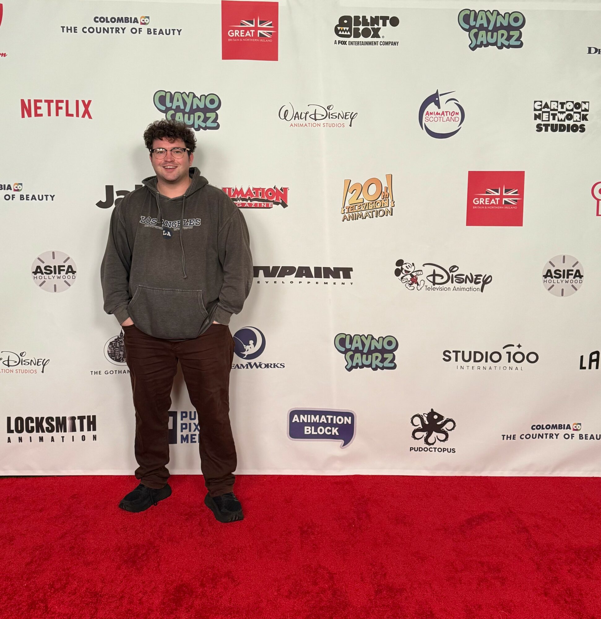 Young person with curly hair and glasses stands on red carpet in front of a backdrop with logos and text.