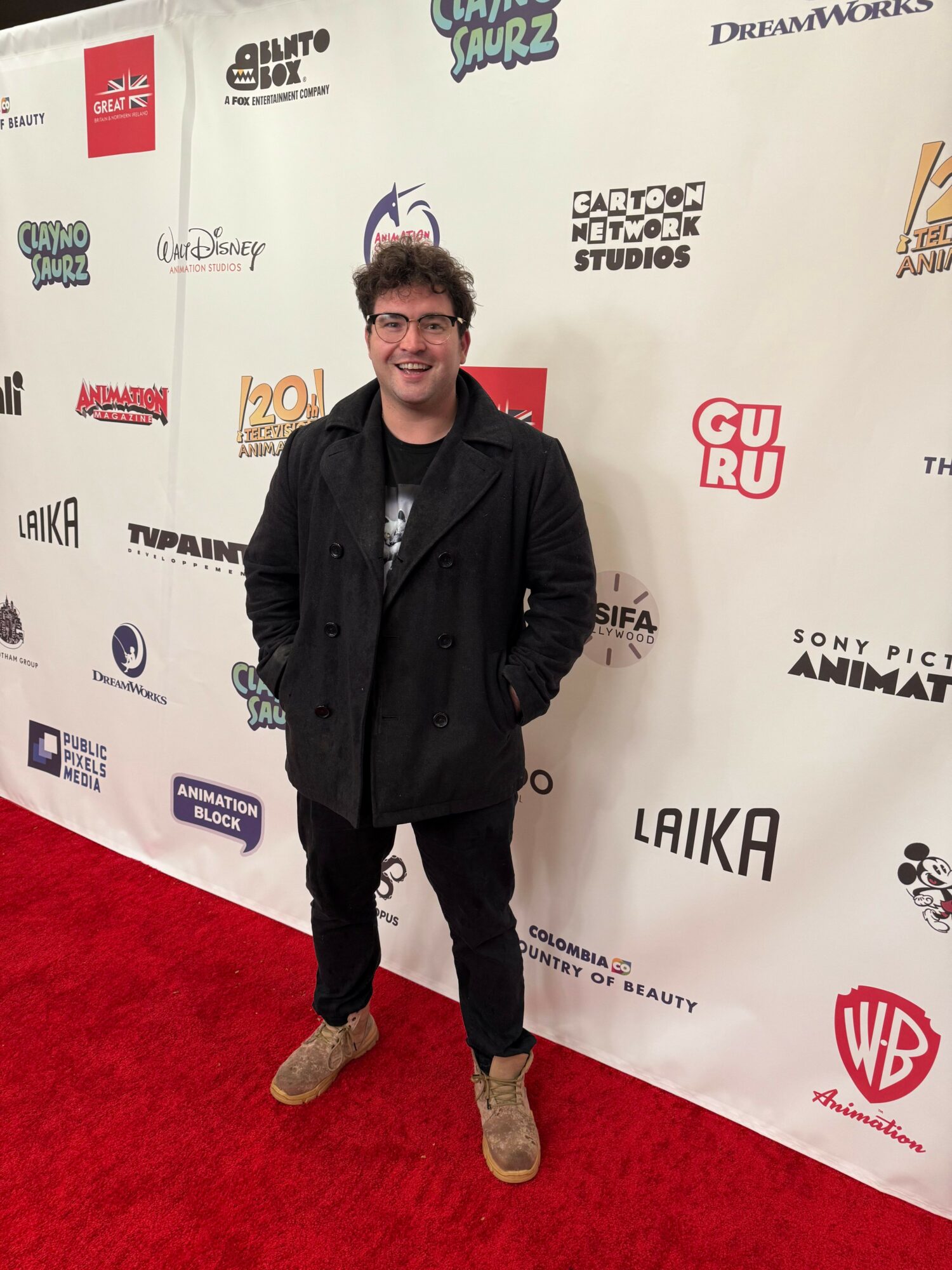 Man in black jacket and pants standing on red carpet in front of a white backdrop with logos.