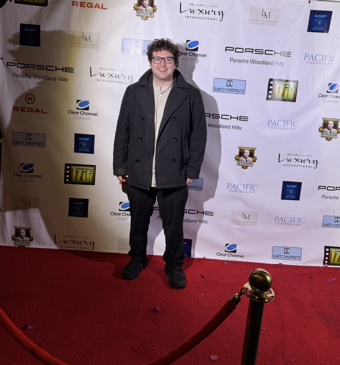 Person with curly hair and glasses standing on red carpet in front of a step-and-repeat backdrop, smiling.