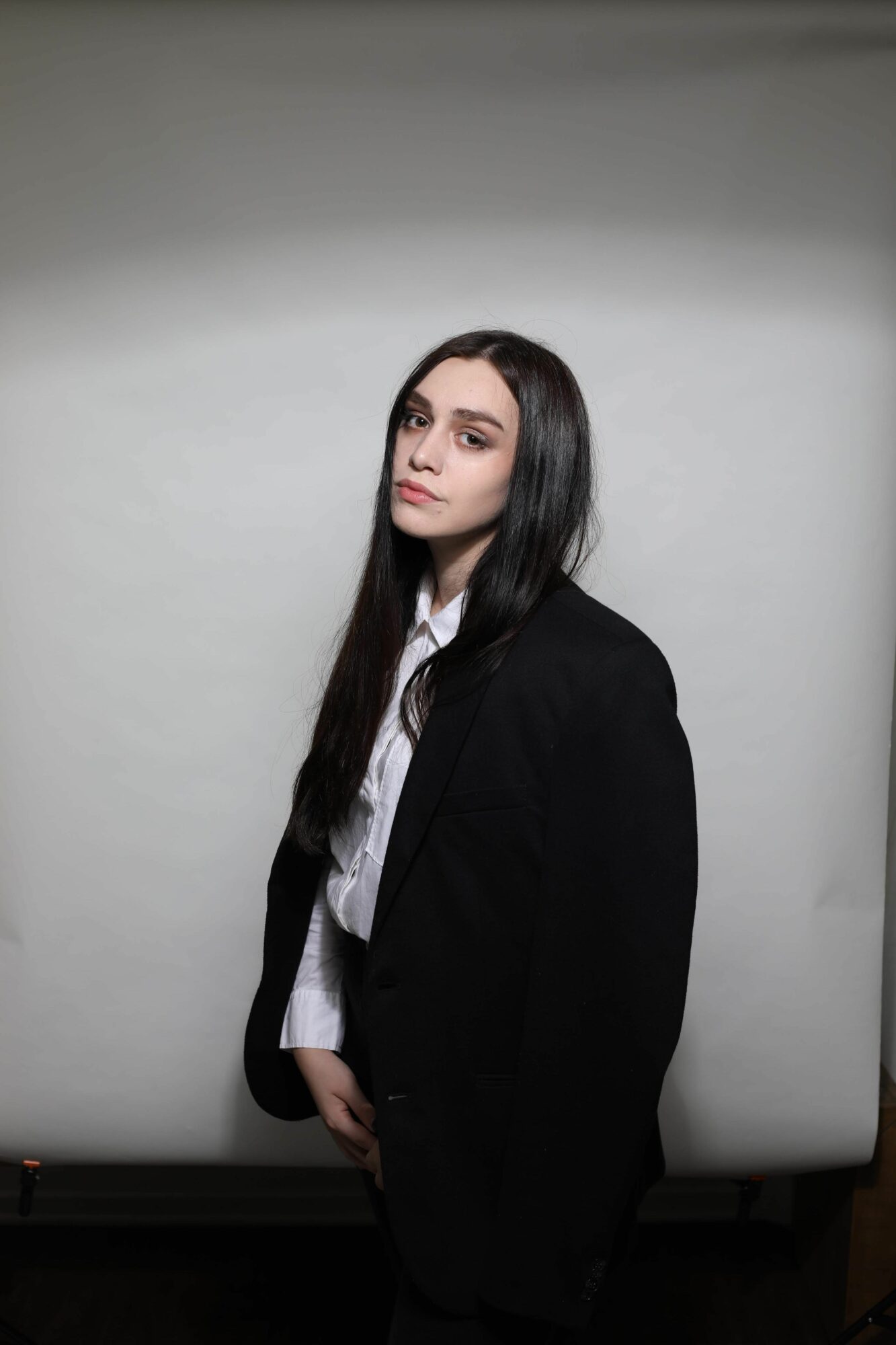 A woman with long dark hair stands sideways against a plain white wall, wearing a black blazer and white shirt.